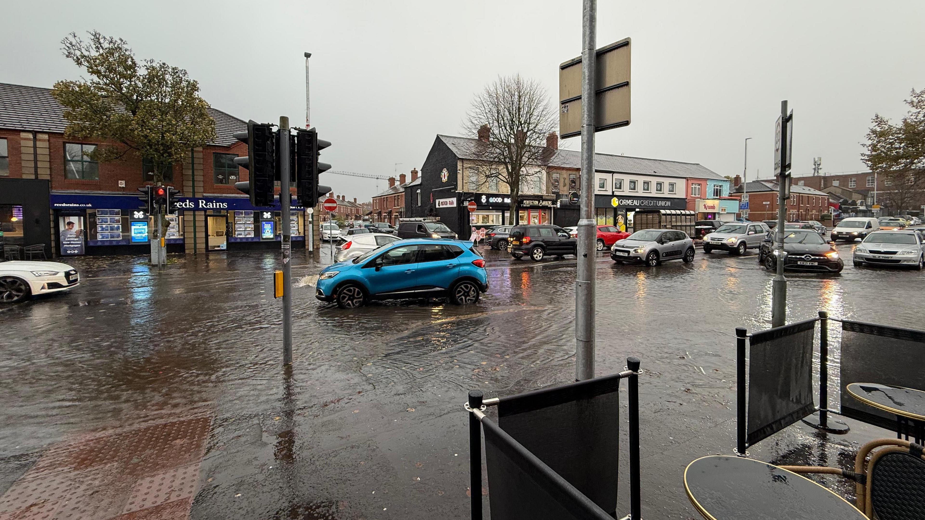 Flooding on a busy high street. A number of cars are on the road.