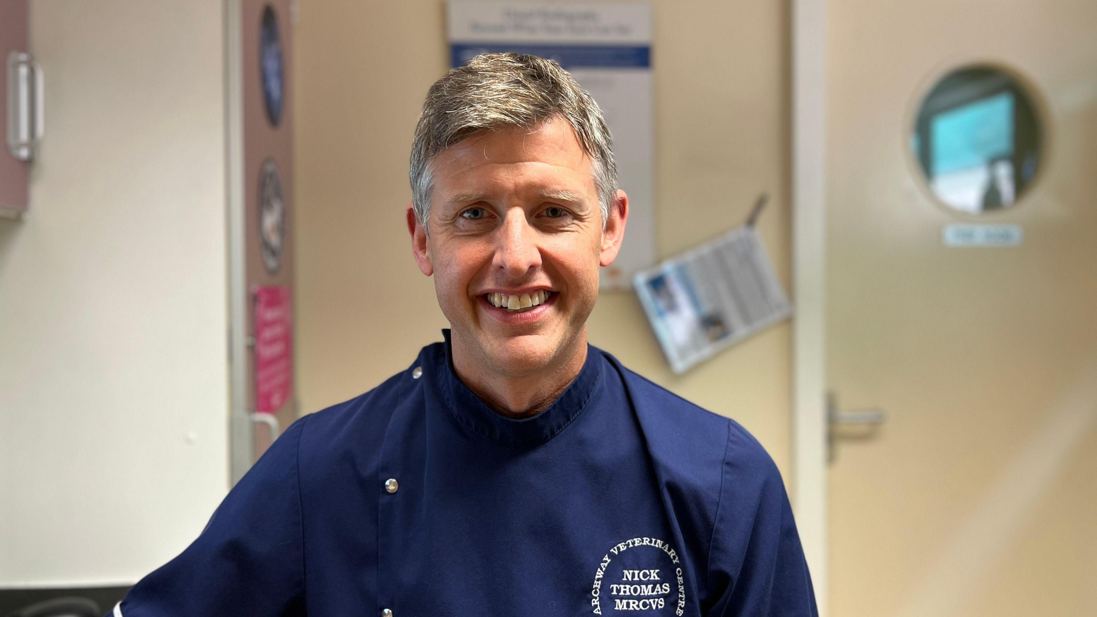 Dr Nick Thomas is a man with grey hair, smiling into the camera. He is wearing navy blue scrubs. Behind him, the cream walls of a veterinary surgery are out of focus. 