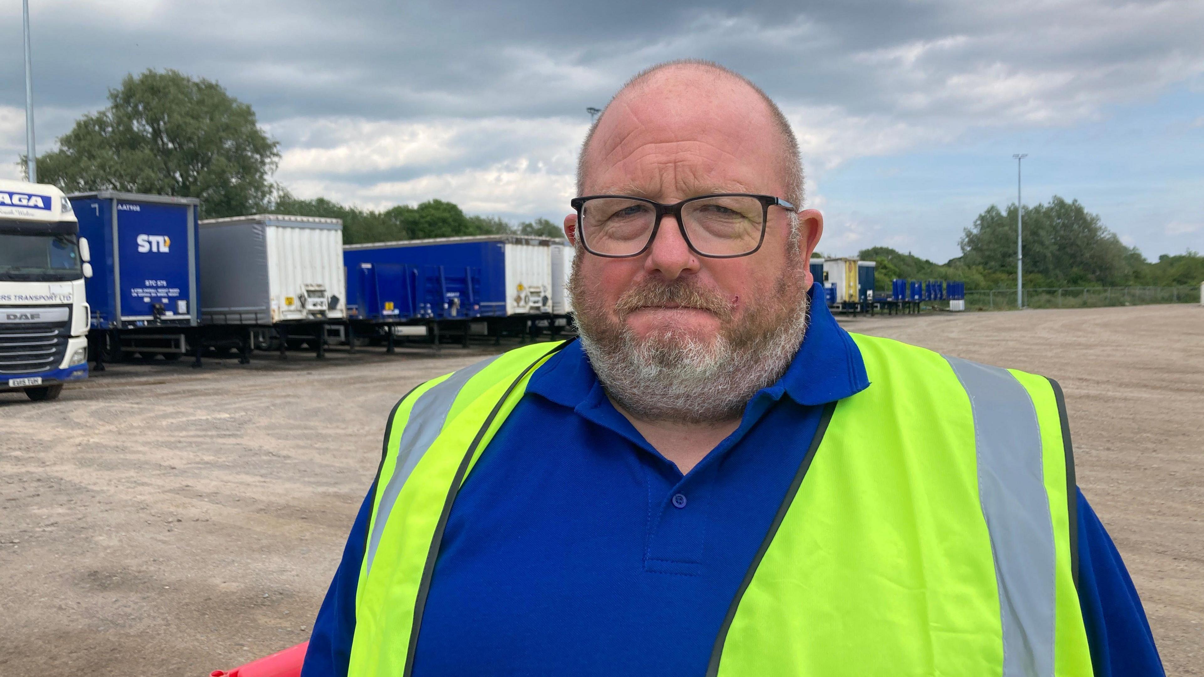 Darren is wearing glasses and a blue shirt with a high-vis vest. Behind him is a row of trucks and trailers which are blue and white. He has a shaved head and a short beard.