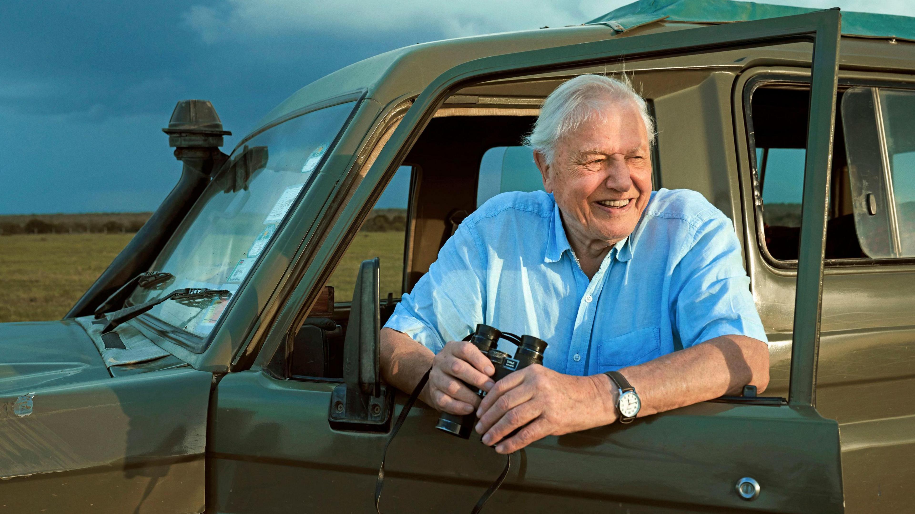 David Attenborough with a pair of binoculars wearing a blue shirt, standing next to a car.