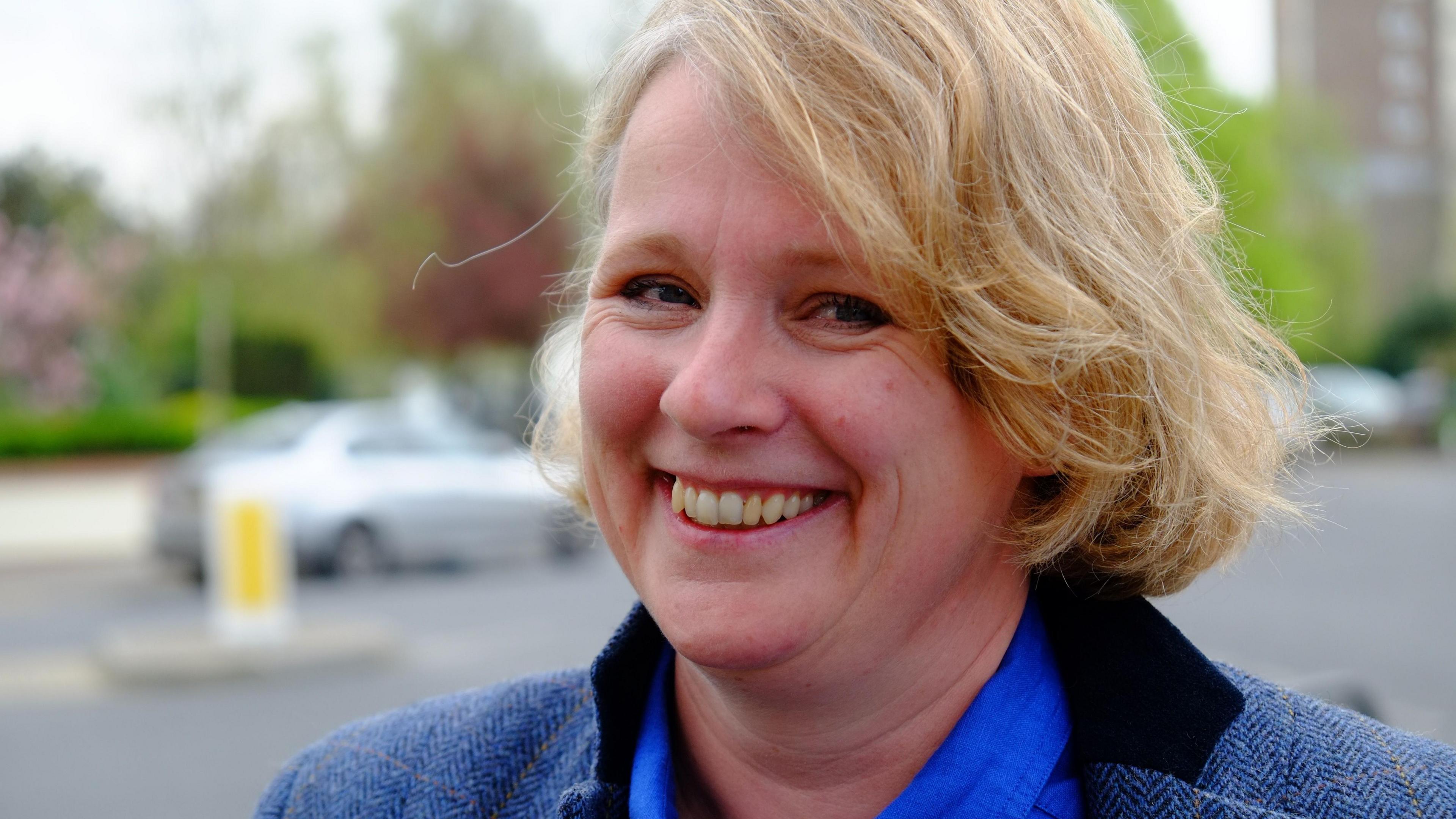 Vicky Ford, the former MP for Chelmsford, smiling at the camera. She is stood by the side of a road with traffic behind her. She is wearing a blue shirt and blue tweed jacket.