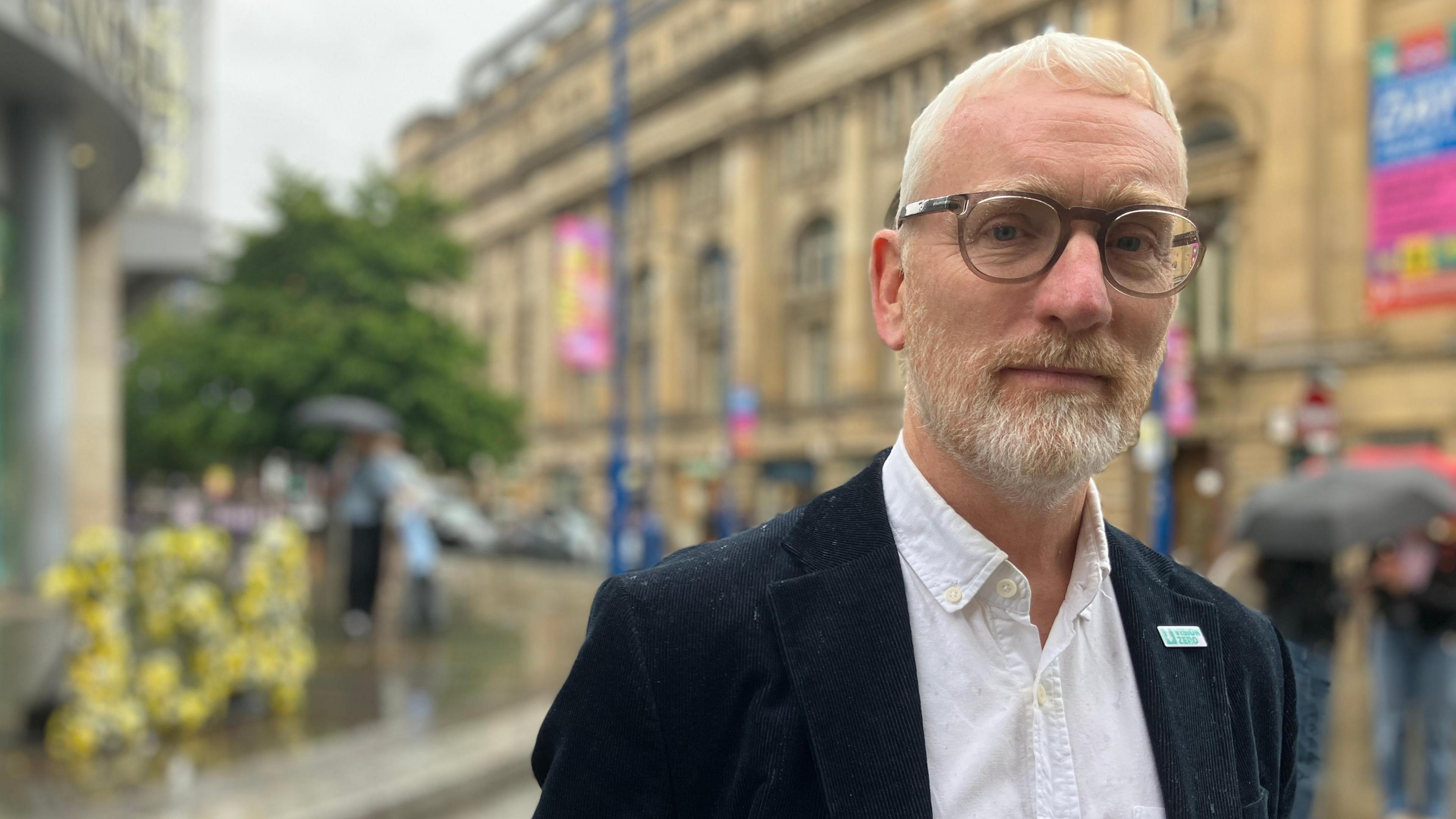 A close up of Peter Boulton. He has short blonde hair, is wearing glasses, and has a short beard. He is wearing a dark jacket and white shirt. In the background is the flower display.