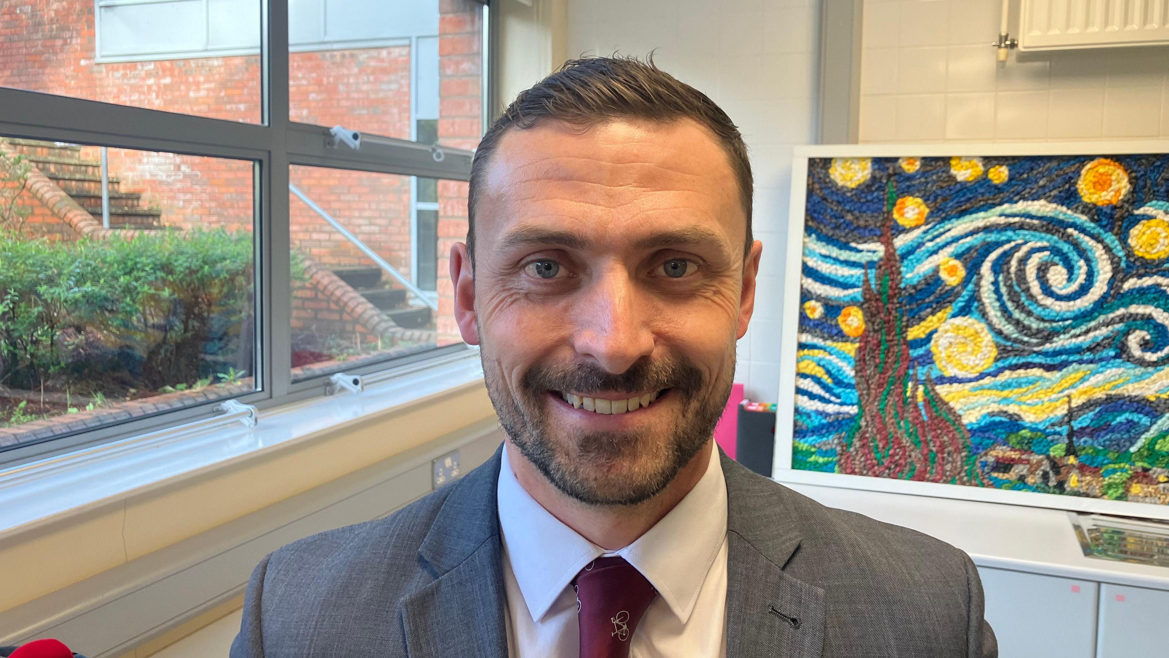 A man in a grey suit, white shirt and maroon tie, stands in front of a window and a painting resembling Van Gogh's The Starry Night.