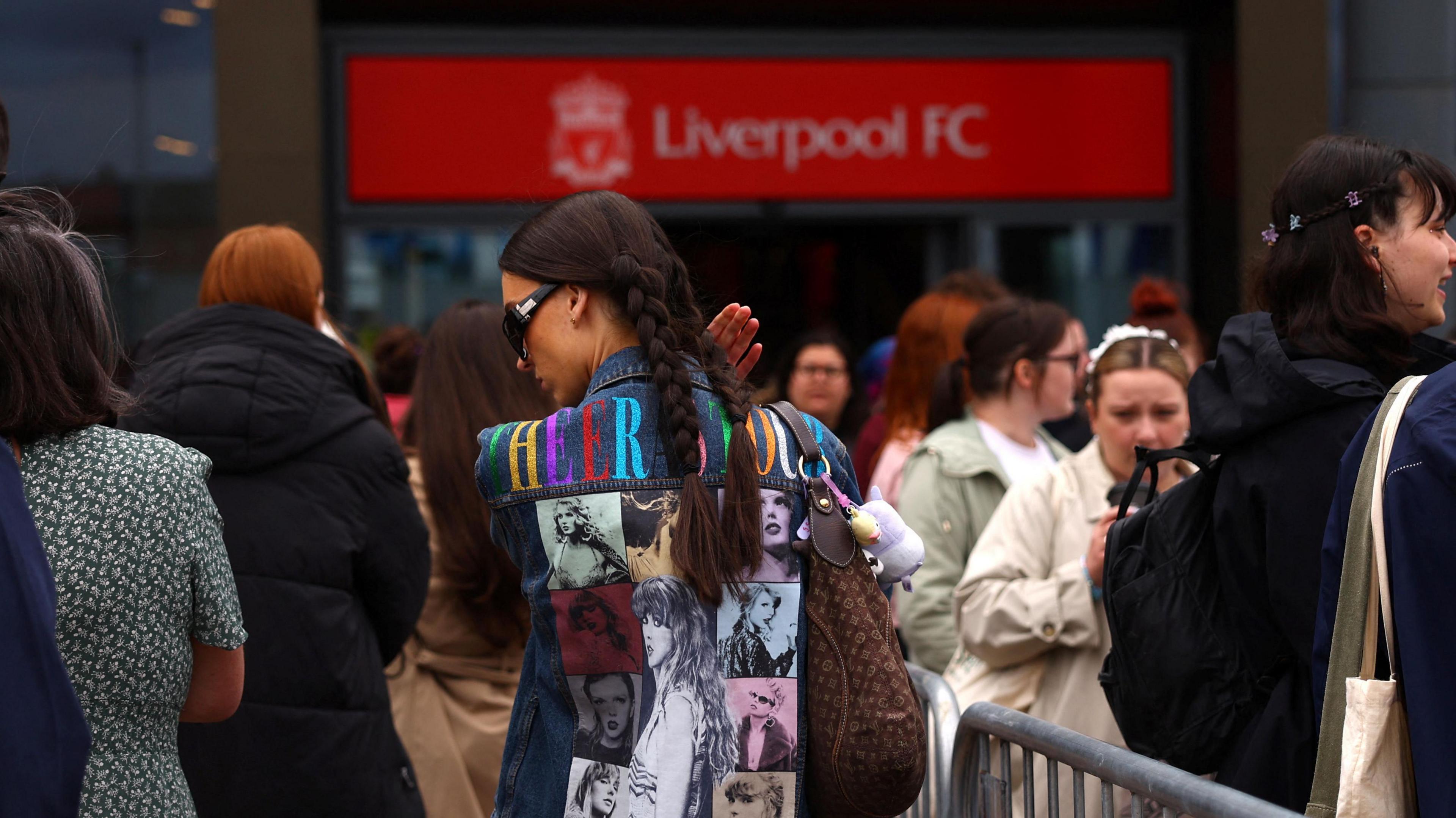 Woman wearing a jacket emblazoned with images of Taylor Swift in a queue snaking past Liverpool FC's store before the Anfield concert