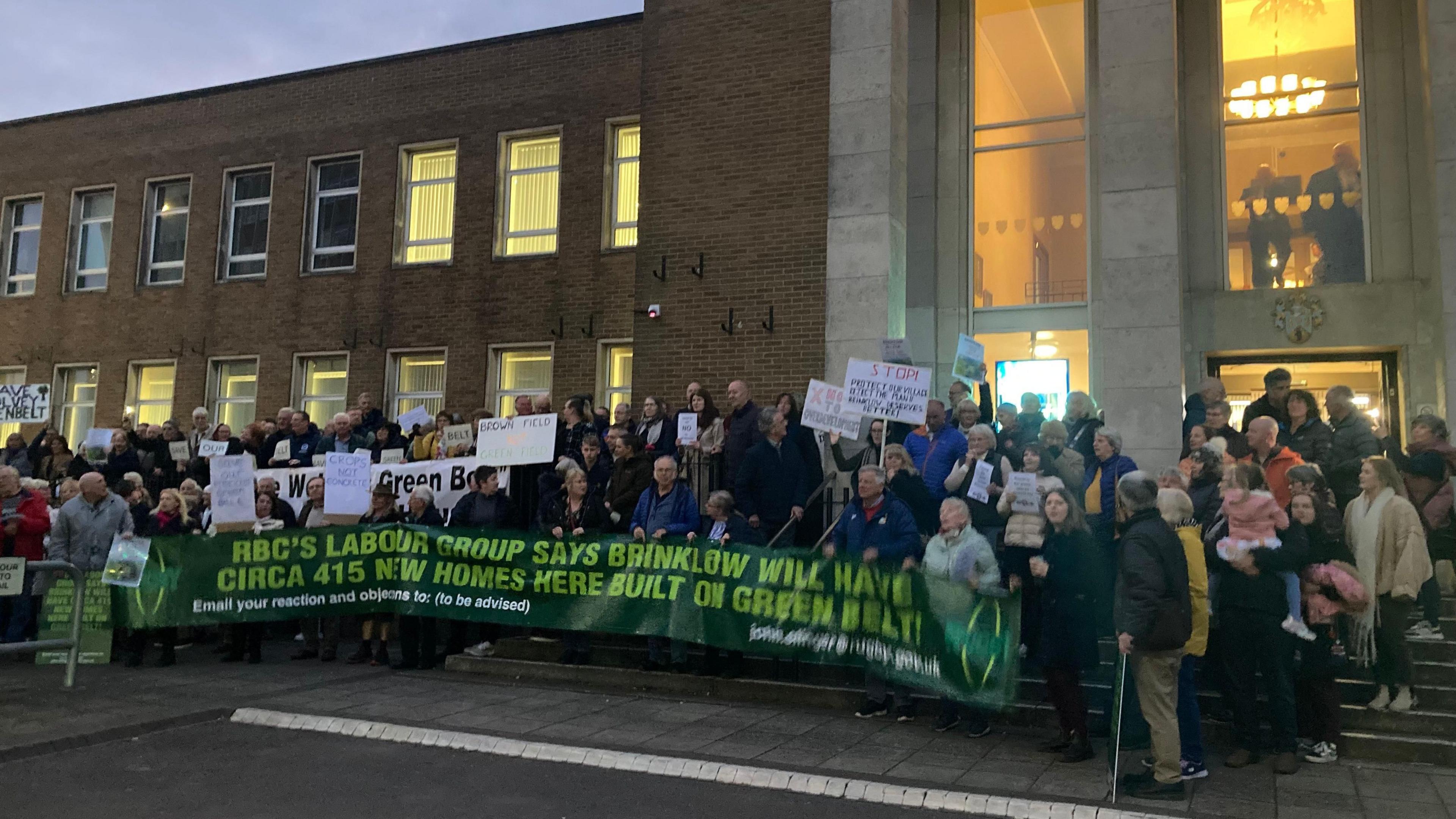 Protesters line up outside Rugby Town Hall which is a two-story brick building attached to a glass and stone two-story entrance. They are holding posters and a banner. The lights are on in the building and it's getting dark outside.