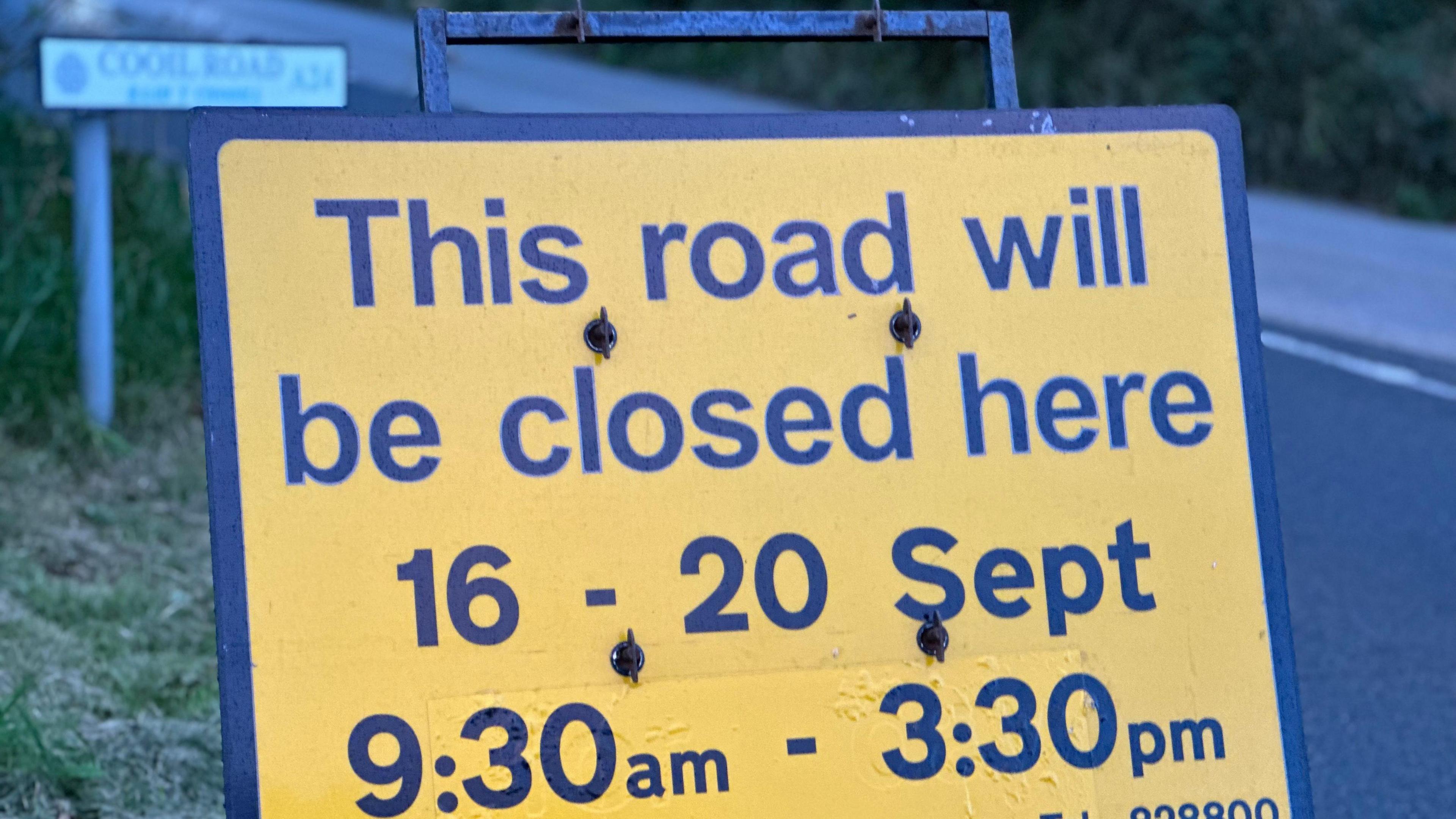 A yellow sign with black writing placed at the side of Cooil Road. It reads "This road will be closed here 16 - 20 Sept 9:30an - 3:30pm.