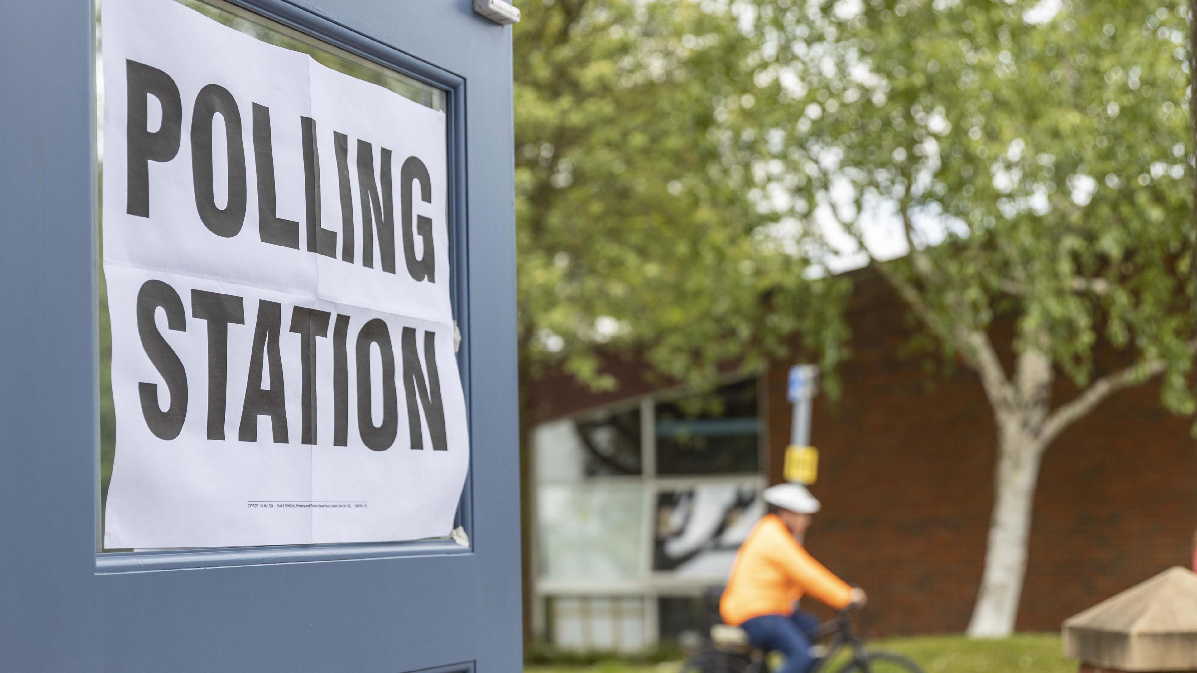 Sign on door saying poling station. A man wearing an orange jacket, jeans and a white helmet is cycling past. 