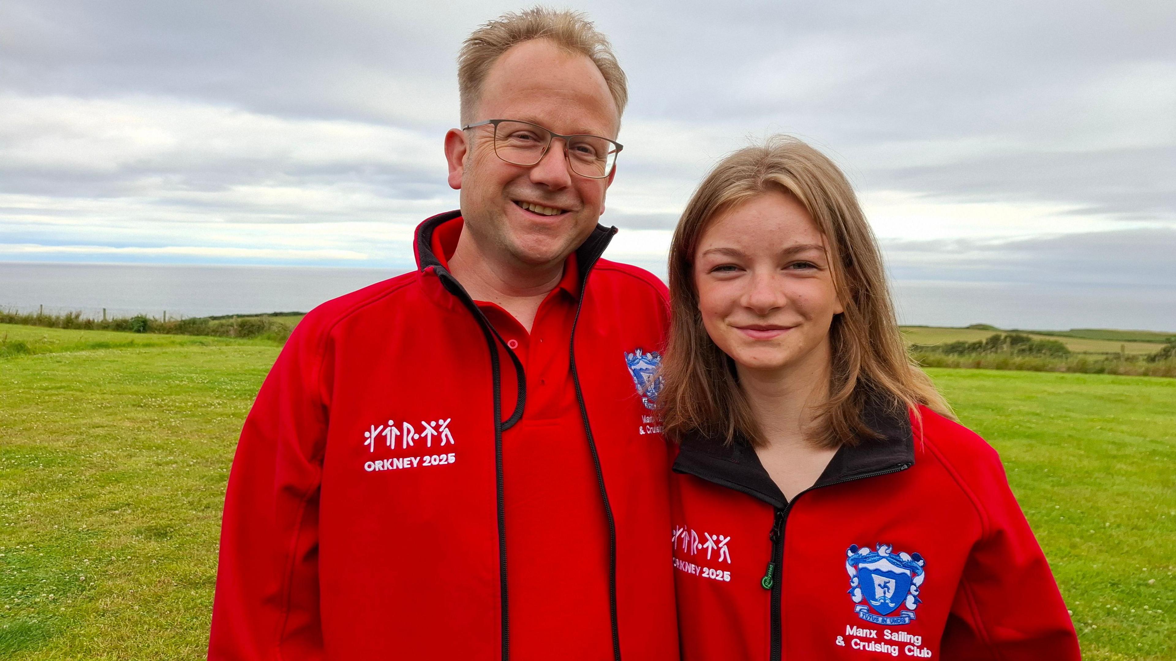 Jason and Lettie Hyett wear red team jackets and stand in a green field. Jason has short fair hair and wears glasses, Lettie has mid-length fair hair.