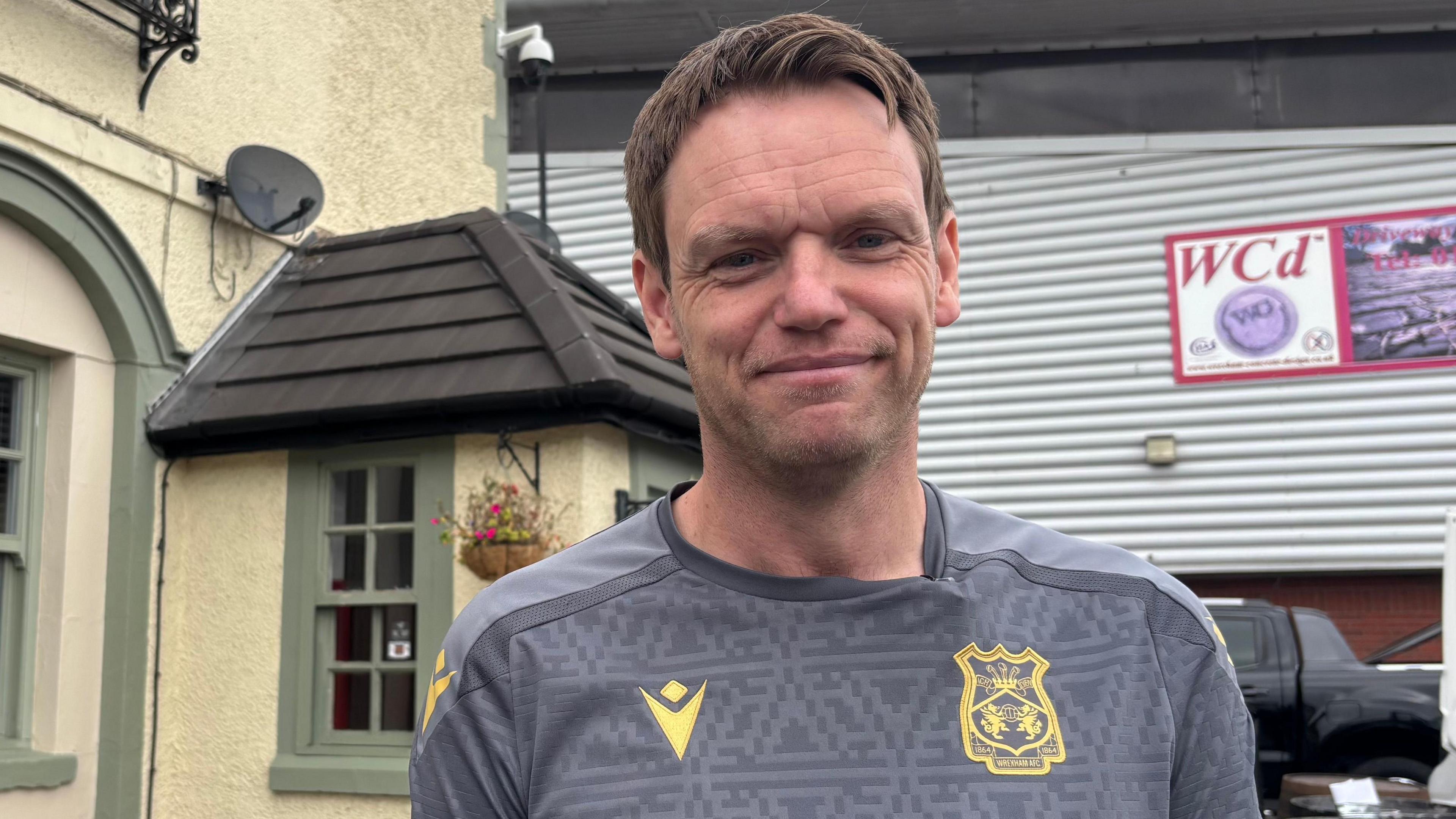Wayne Jones smiling at the camera. He is stood outside the Turf Hotel which stands next to the Racecourse stadium. he has a grey Wrexham shirt on with gold embroidery. He has short, dark hair. Behind is the pub and the Wrexham football ground.