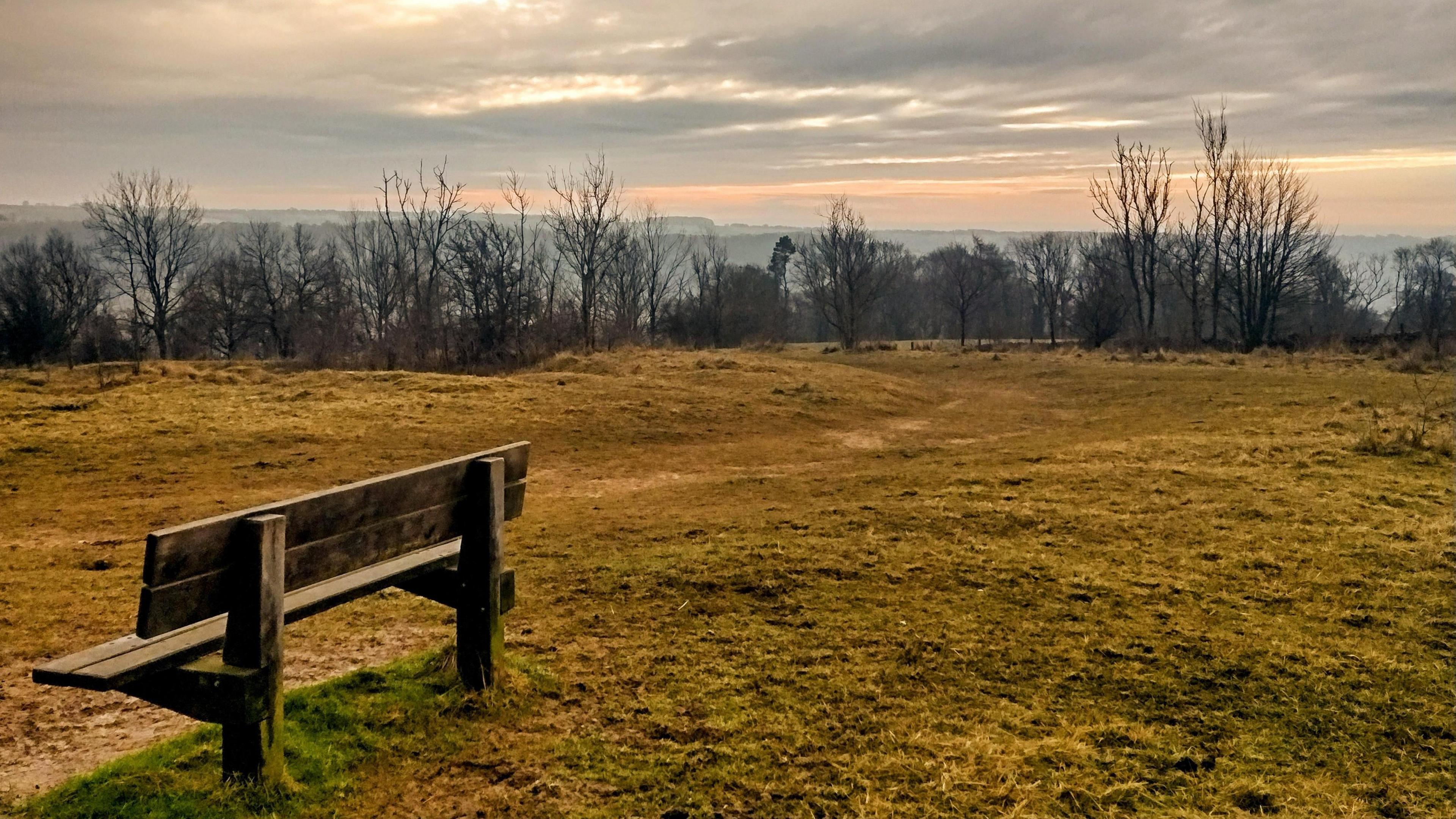 A bench in the forefront of the picture with a grey sky and the sun creeping through the clouds.