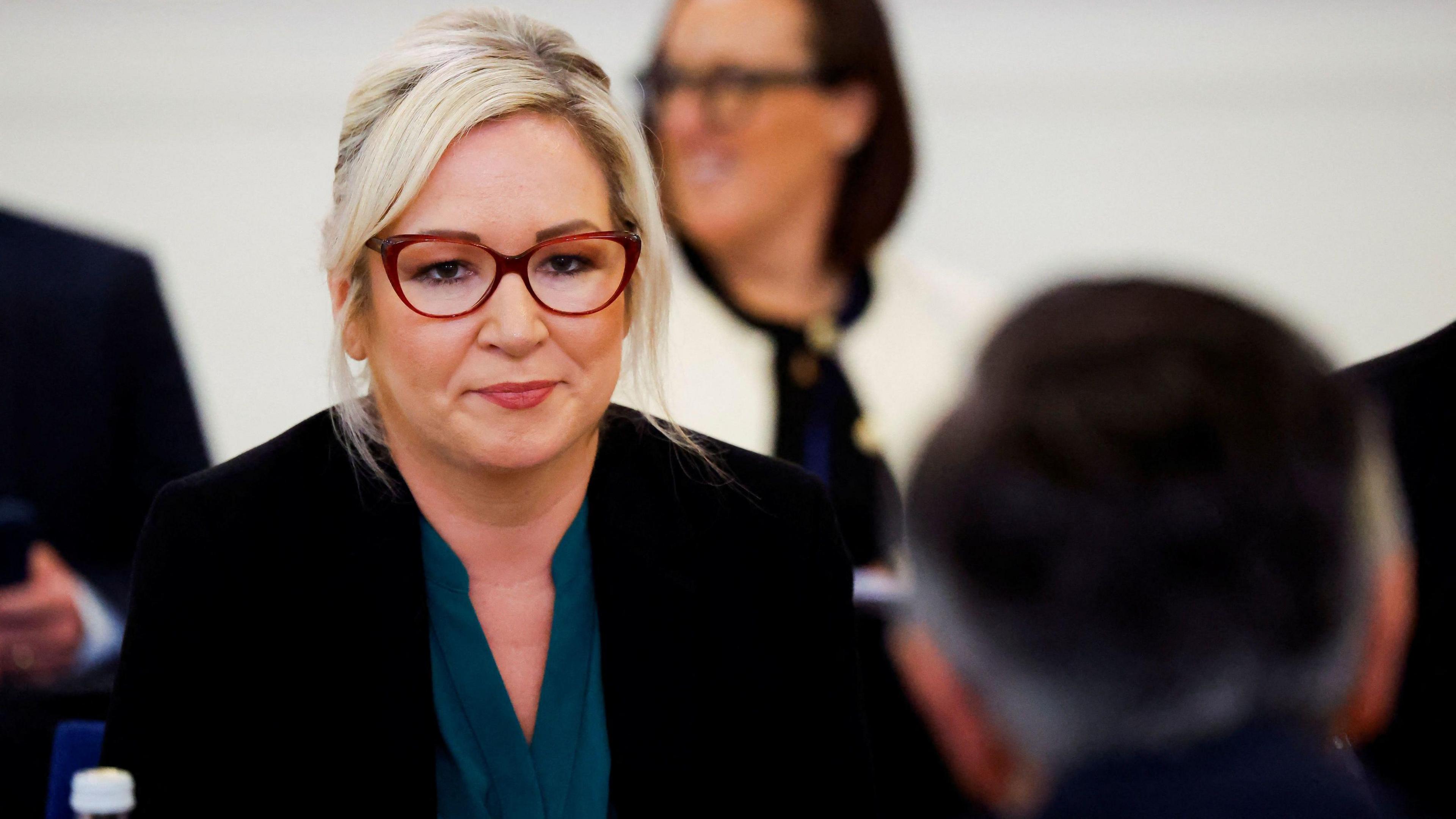 Michelle O'Neill - a woman with blonde hair and red-rimmed glasses - looks across a table to a man sitting opposite her. Only the back of his head can be seen. She is wearing a black jacket and teal blouse.