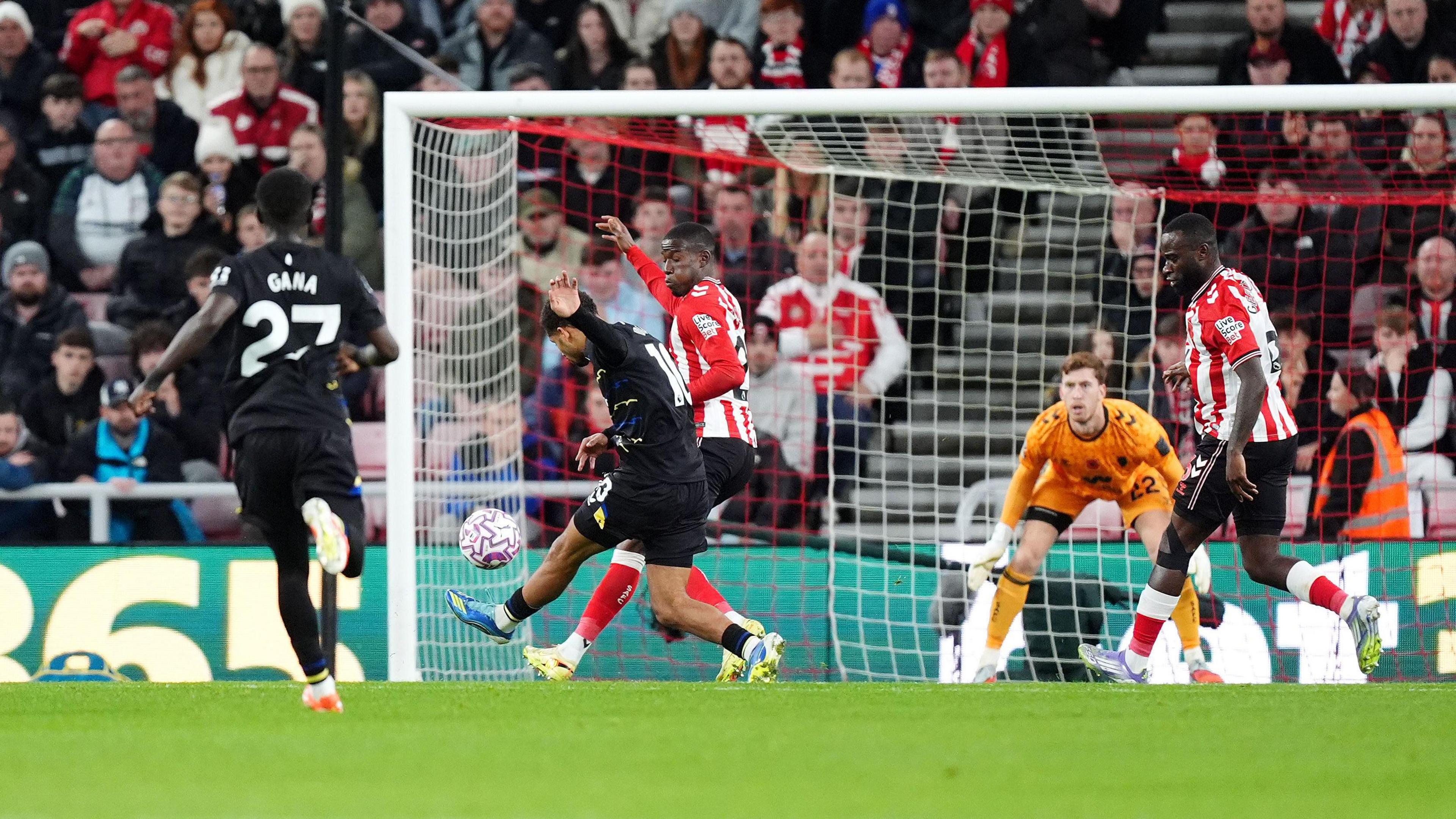 Iliman Ndiaye wears a black kit. He can be seen kicking a ball into a goal on a football pitch as a player in red and white jerseys tries to intercept. They are on a green football pitch and the goal keeper wears an orange jersey.