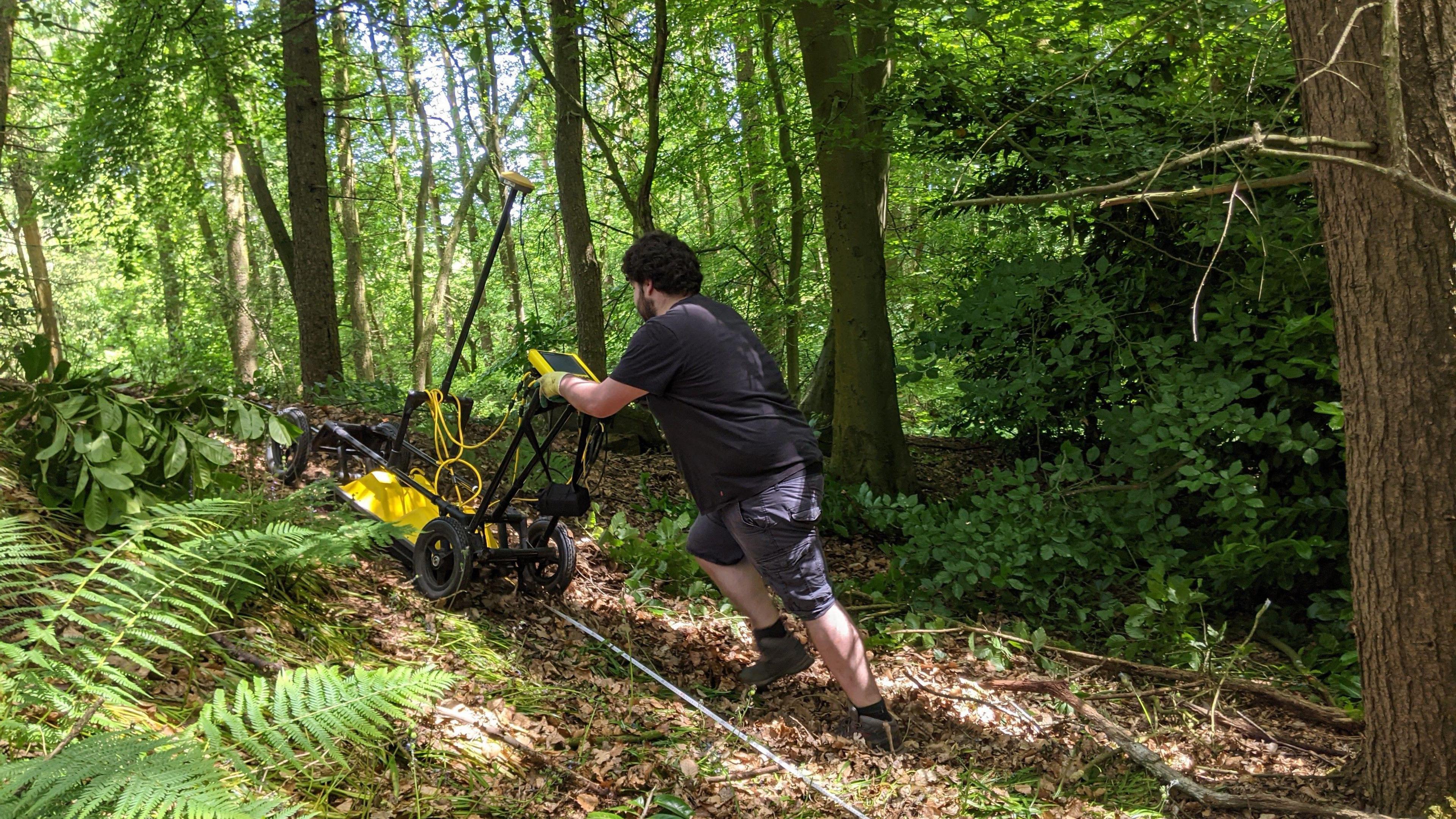 A man in a dark t-shirt and shorts, pushing a piece of equipment that looks like a lawnmower, which is surveying underground. It is yellow and black and has lots of leads coming off it, with a screen on its handle. The area around the man is woodland with green trees and leaves.