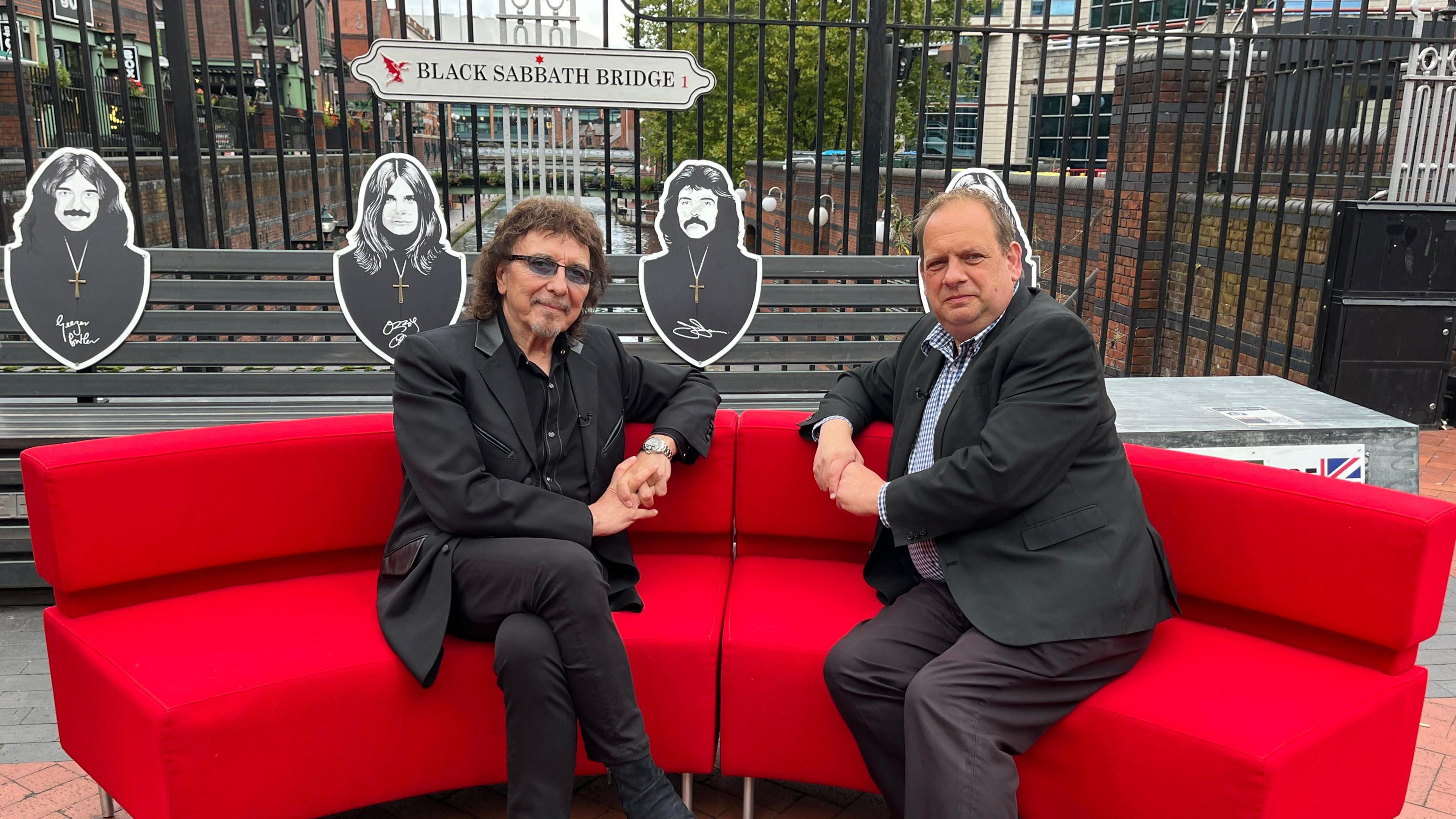 Tony Iommi sits on the red sofa, which is placed in front of the Black Sabbath bench on Broad Street in Birmingham. Sitting next to him is reporter Ben Sidwell