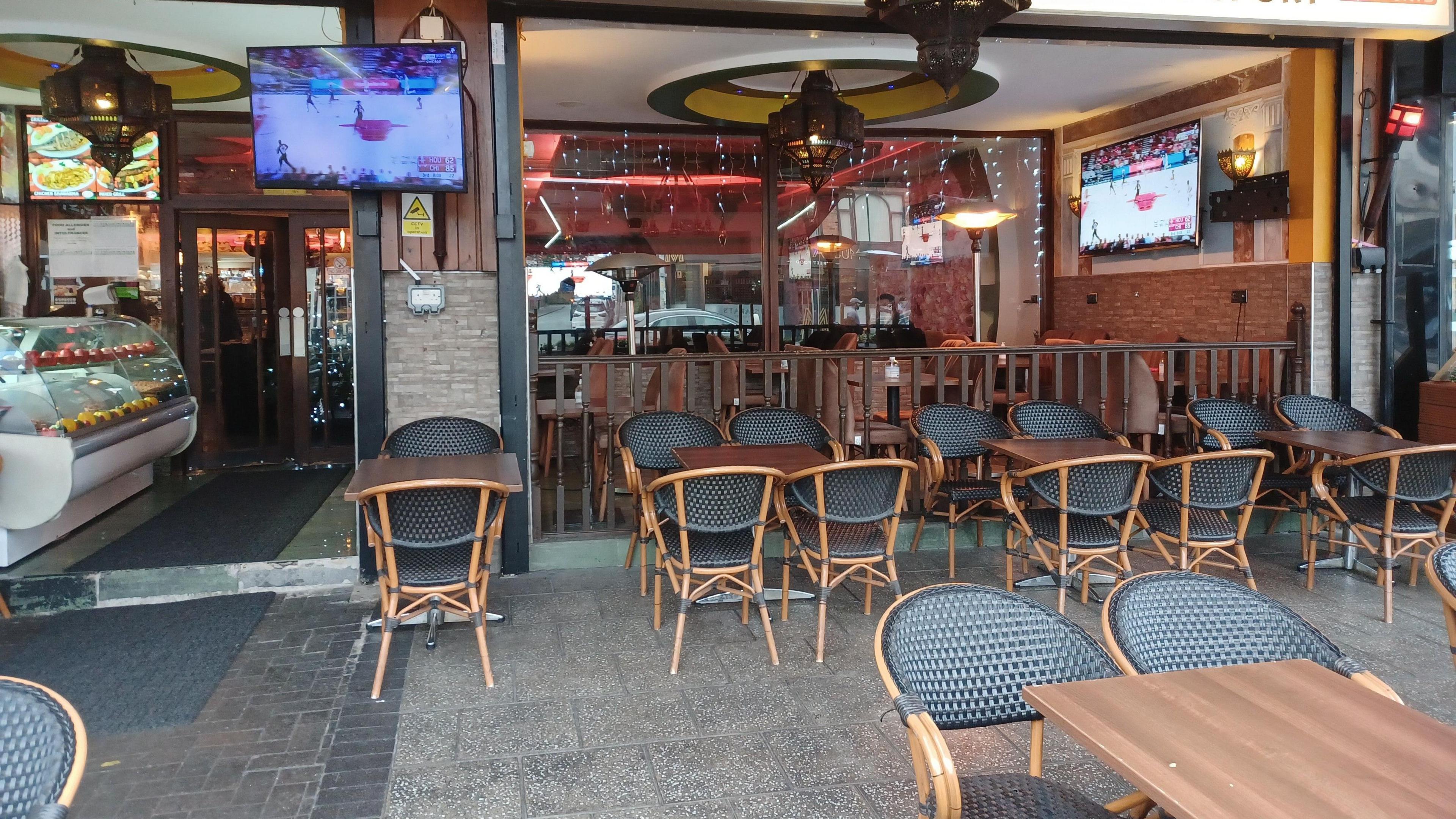 Chairs and tables lined up outside of the opened up front of Antalya cafe, with a display counter to one side, and TV screens placed on the walls.