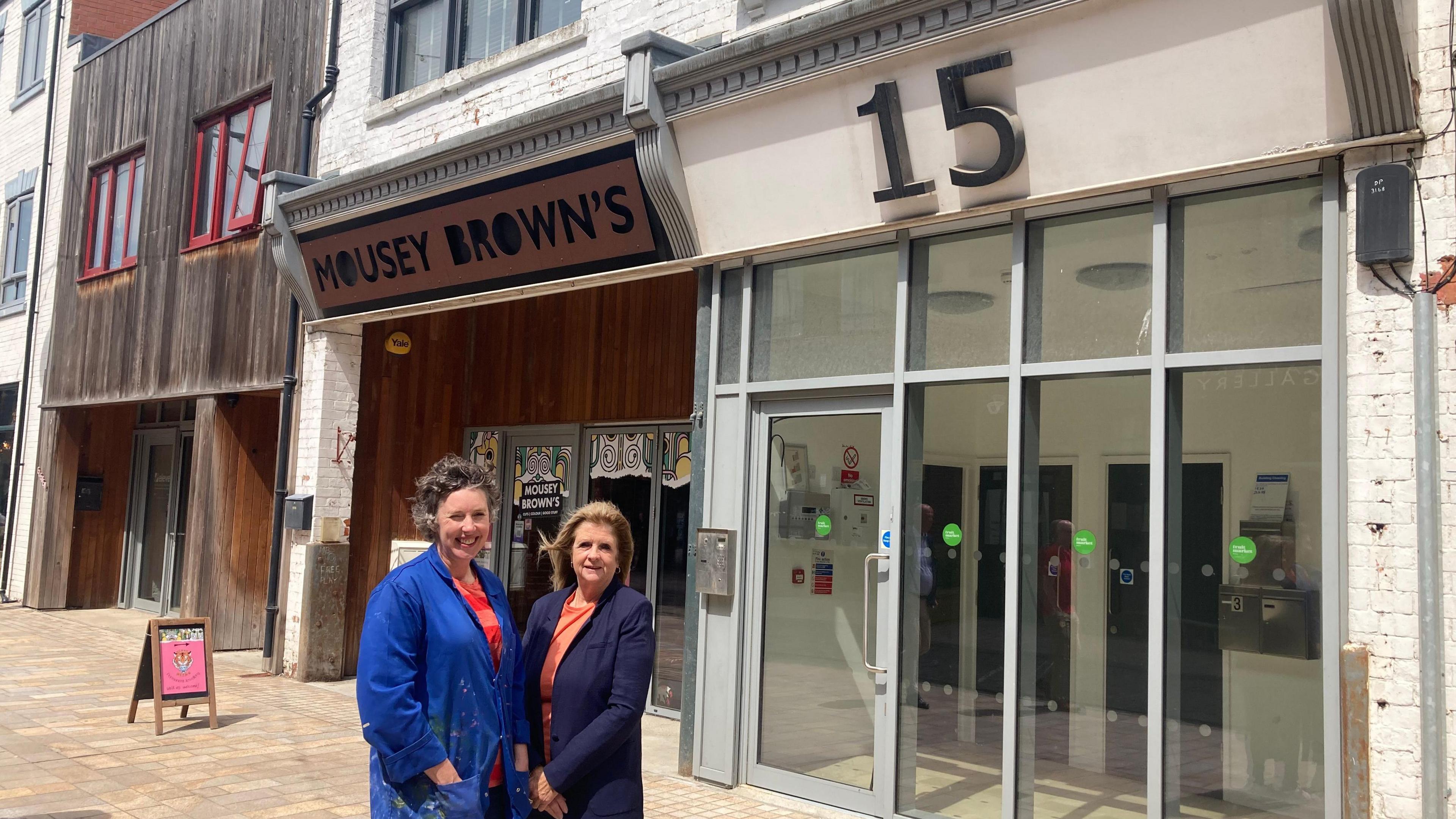 Lady smiling with short wavy hair, blue denim jacket and striped red and orange jumper with lady with long brown hair, blue jacket and orange top.
They are stood on a paved street in front of wood and glass buildings with an advertising board outside.