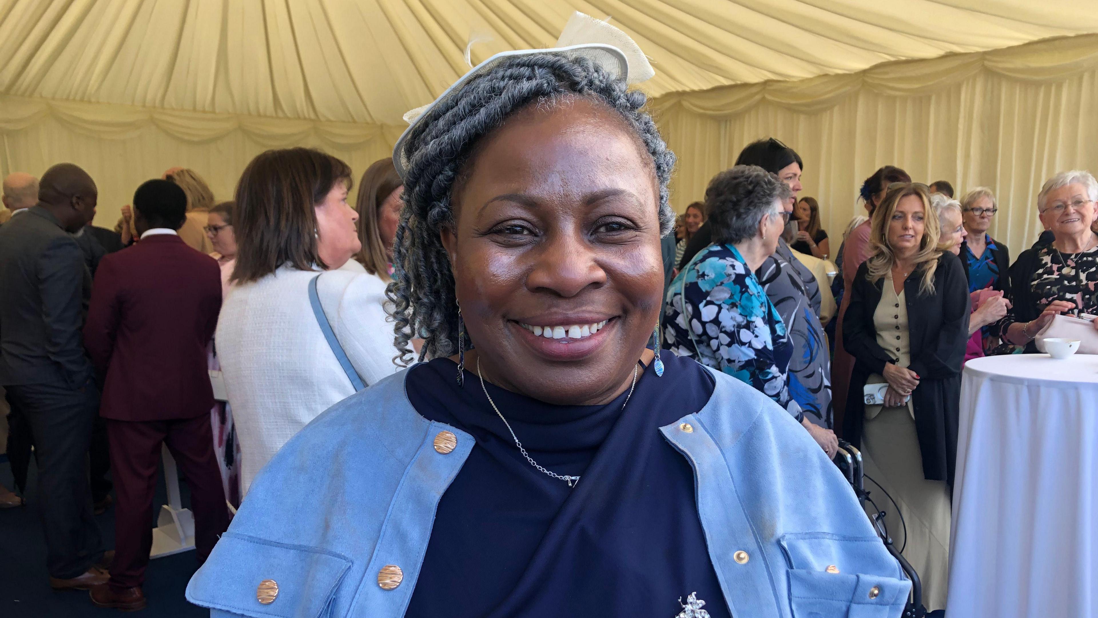 Women standing in navy blue dress and light blue jacket while standing under a garden party tend. She is also wearing a white hat for her head. 