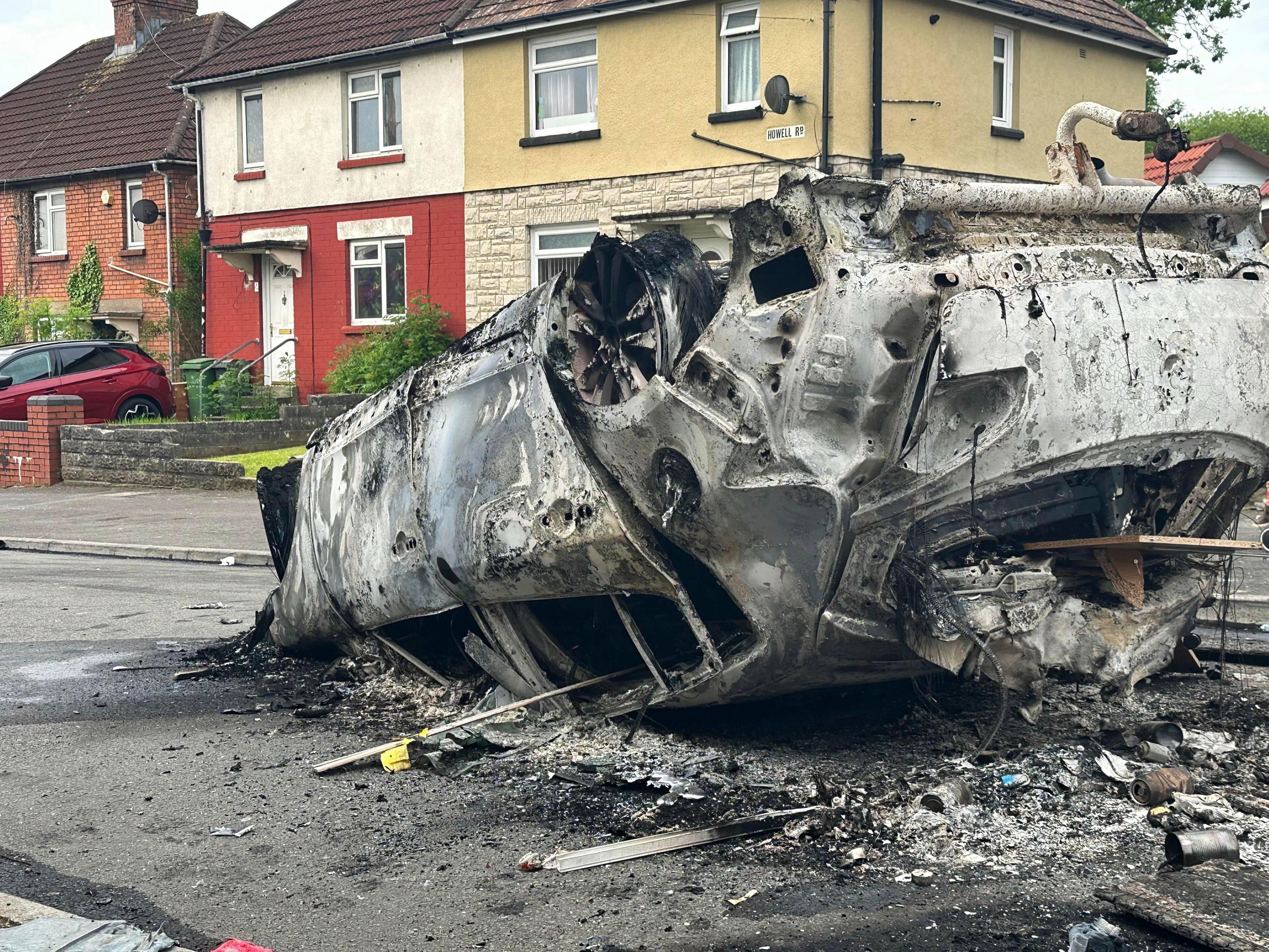 An upturned burnt out car in front of houses. 