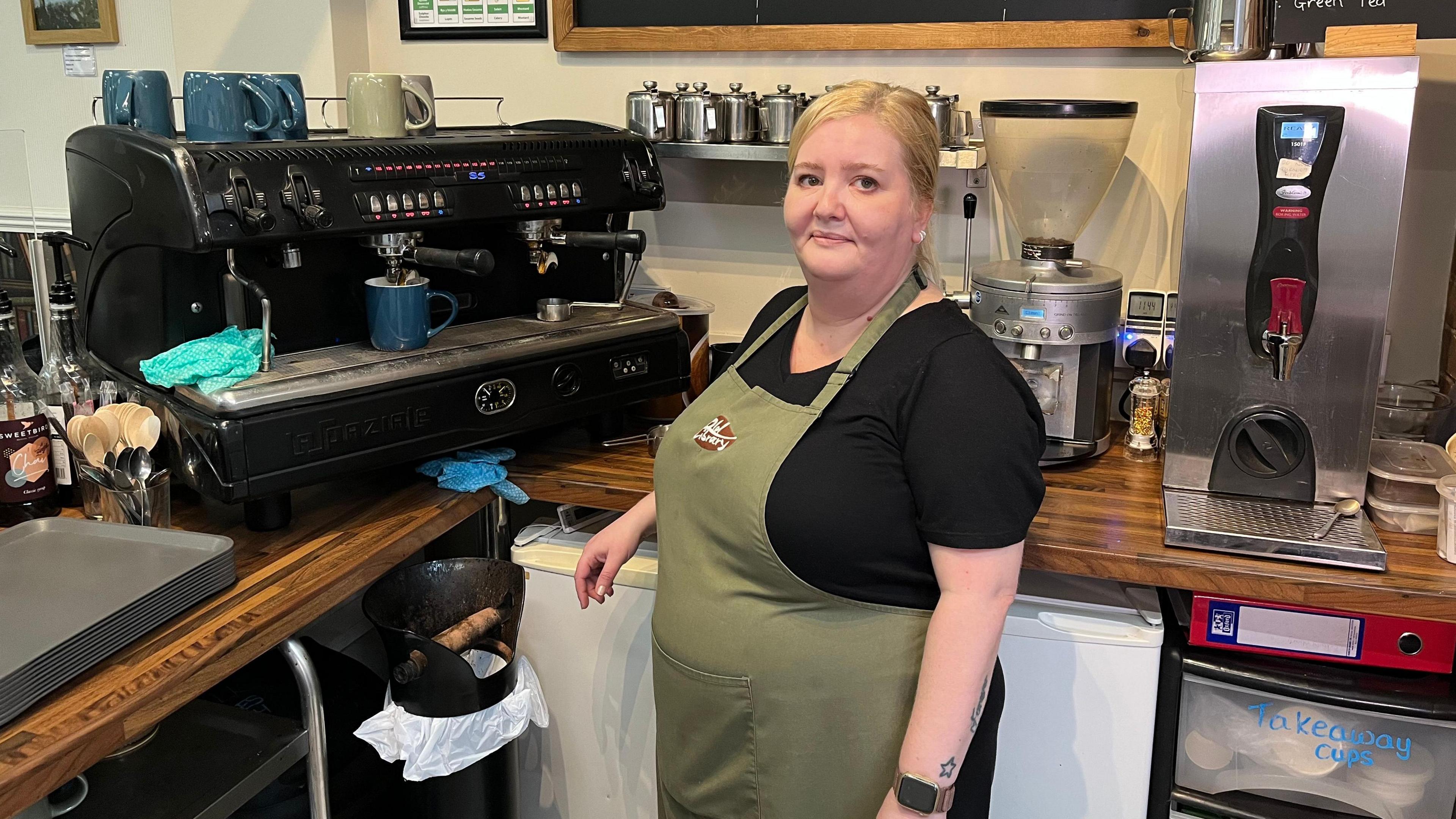 Rachel standing next a large coffee machine in her café. She is wearing green apron over a black top.