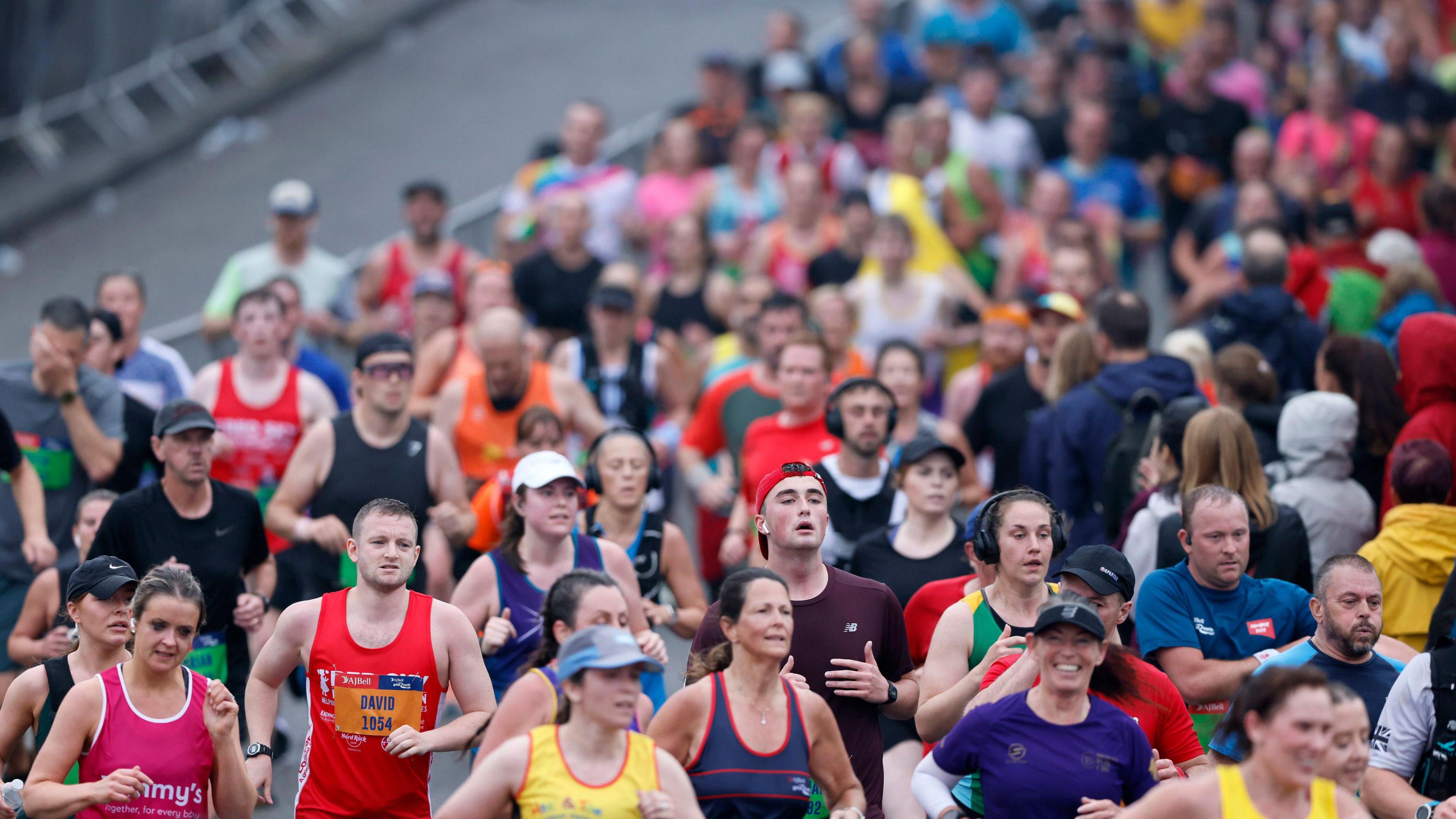 A crowd of runners. They are wearing a variety of coloured tops, with race numbers attached to some. Some wear headphones and others wear caps. One woman smiles at the camera.