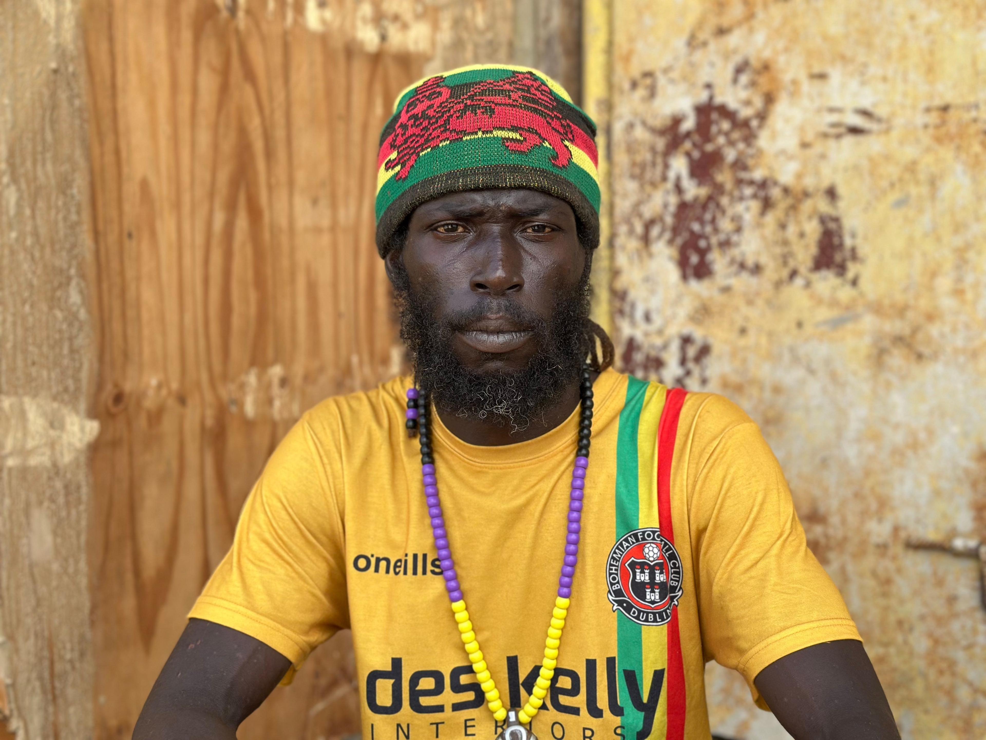 Roy Perry wearing a yellow t-shirt seated in front of a badly damaged building