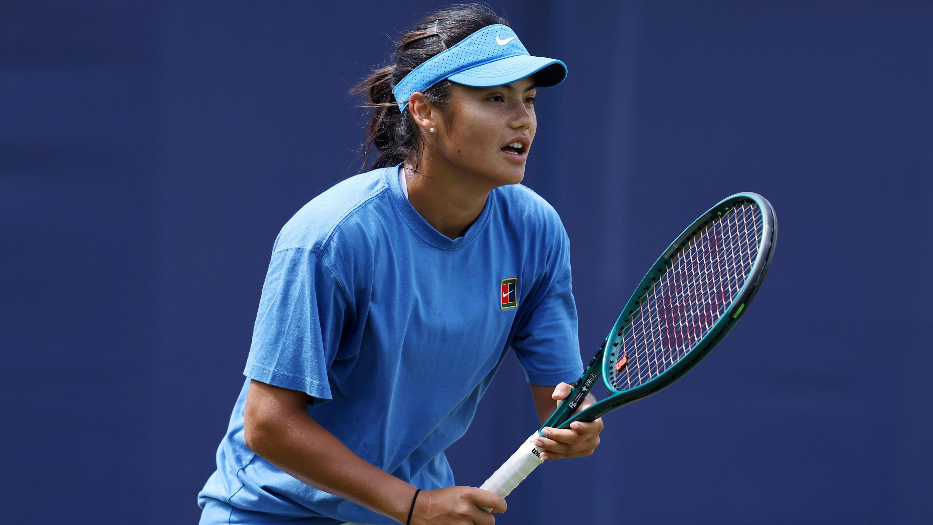 Emma Raducanu is wearing a blue t-shirt and sun visor and holding a tennis racquet.