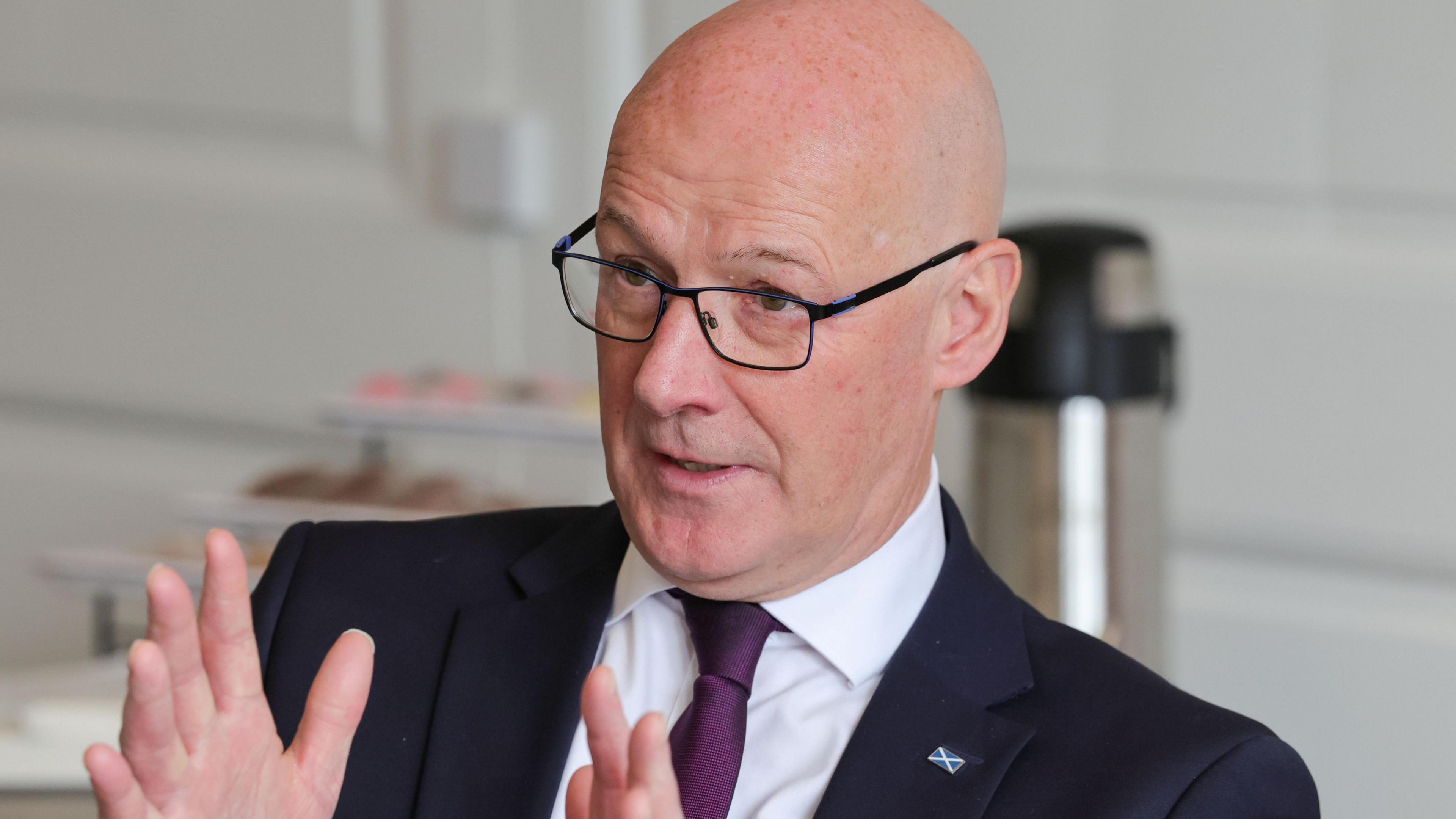 John Swinney wearing a dark suit over a white shirt and a dark tie is holding both of his hands up in front of him