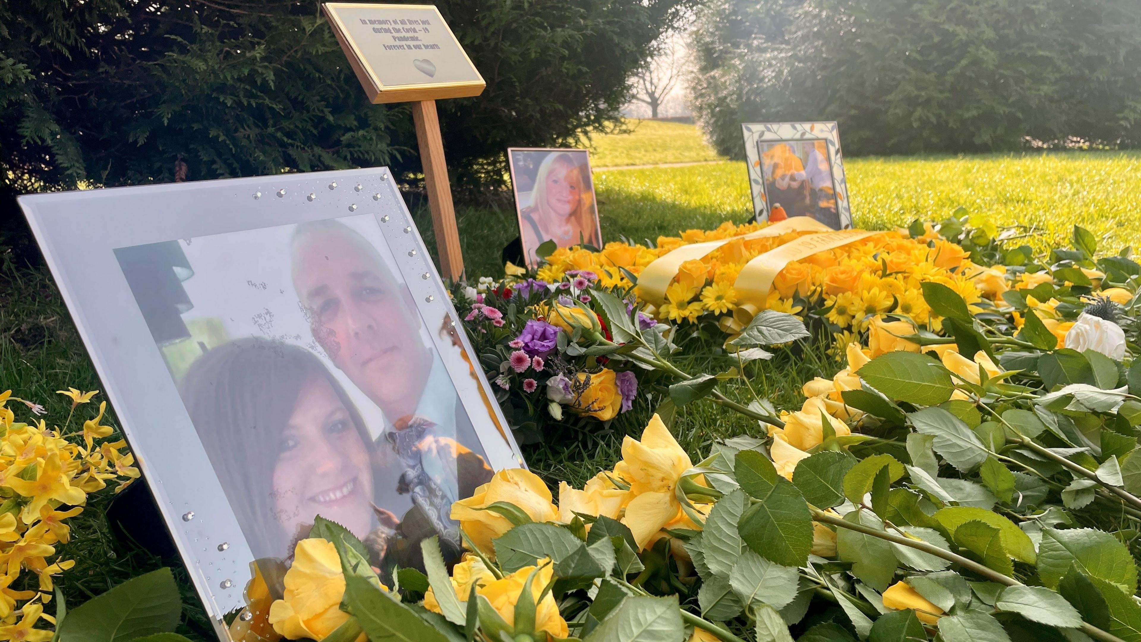 A close up of photos of the covid dead and flowers lying on the ground in a park. The flowers are almost all yellow and sunlight can be seen hitting the ground through trees in the background.