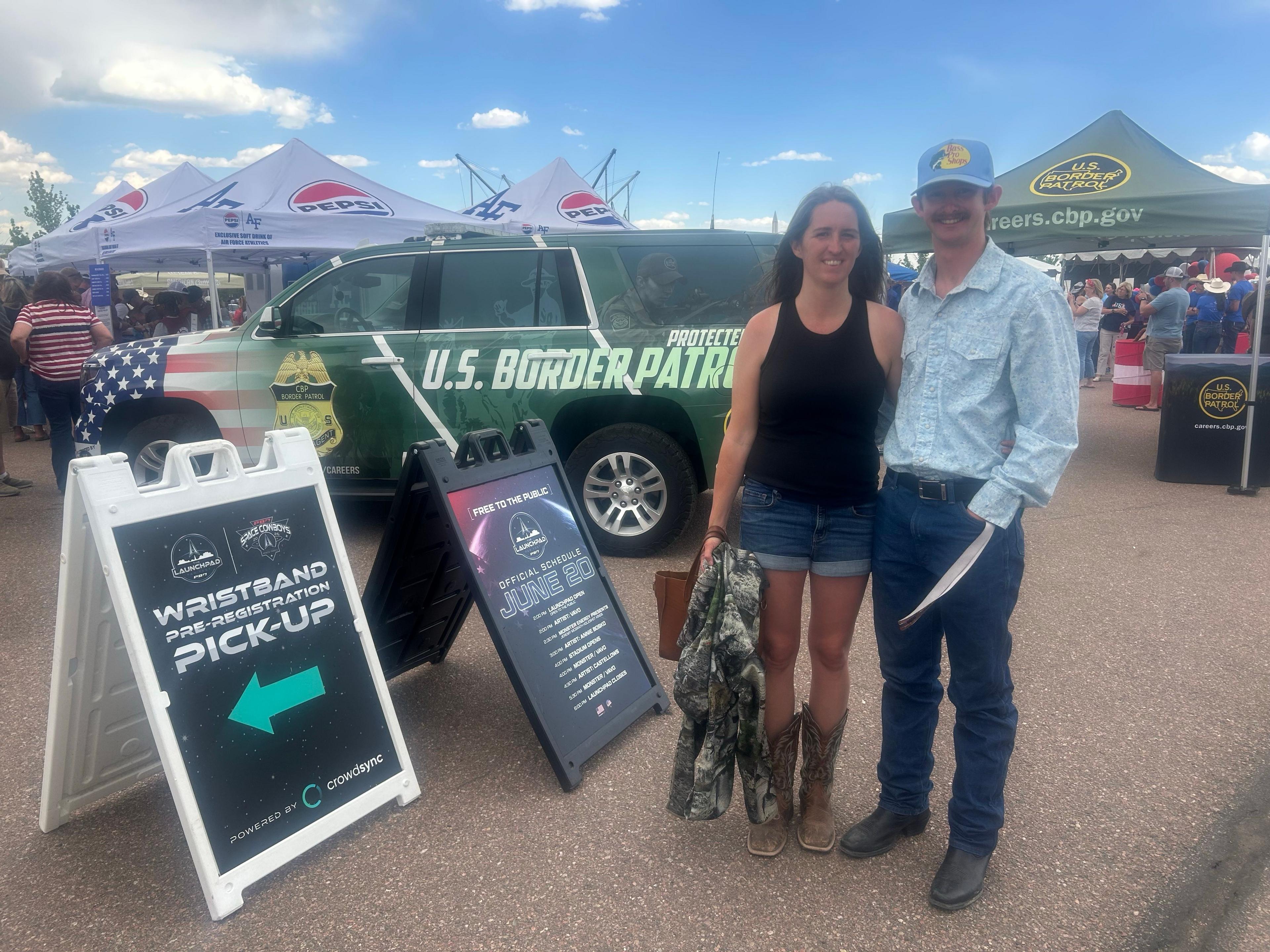 A young man in a baseball hat stands in front of a tent advertising the Customs and Border Patrol, with his mom standing next to him in cowboy boots