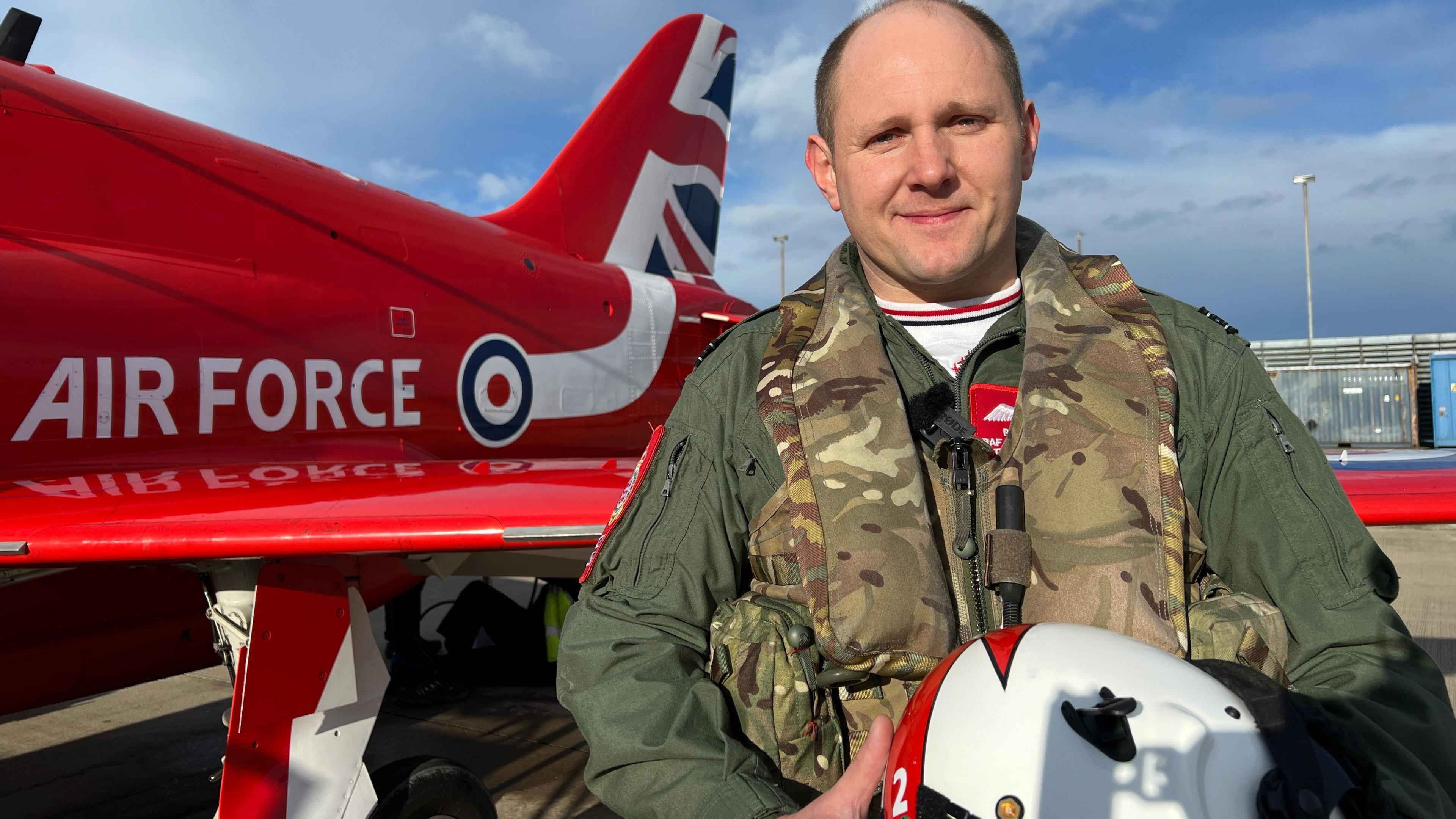 Rob is standing next to the wing of a red arrow aircraft. He's holding his white and red helmet with the number two on it. He's wearing a green jumpsuit and green camouflage jacket on top. Rob is smiling.