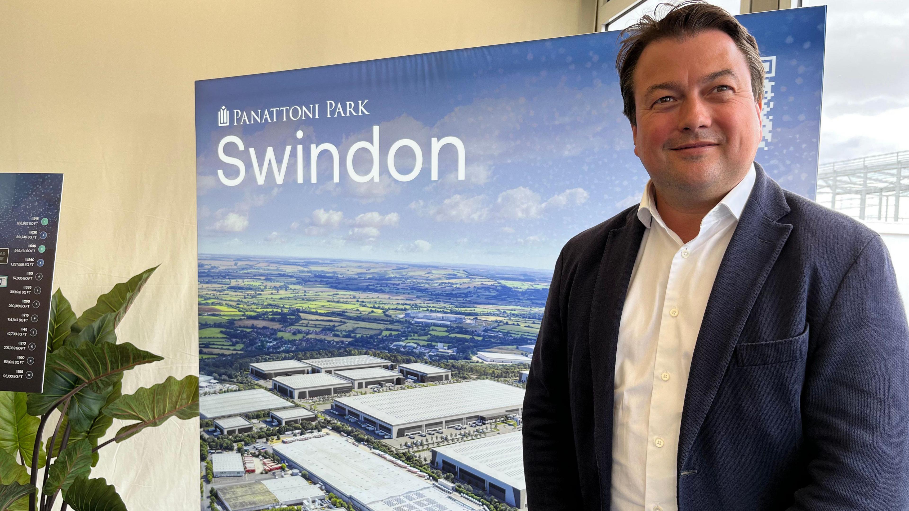 James Watson stands to the right in suit jacket and shirt. He is smiling and stands in front of a large banner with a computer graphic showing what the completed park will look like