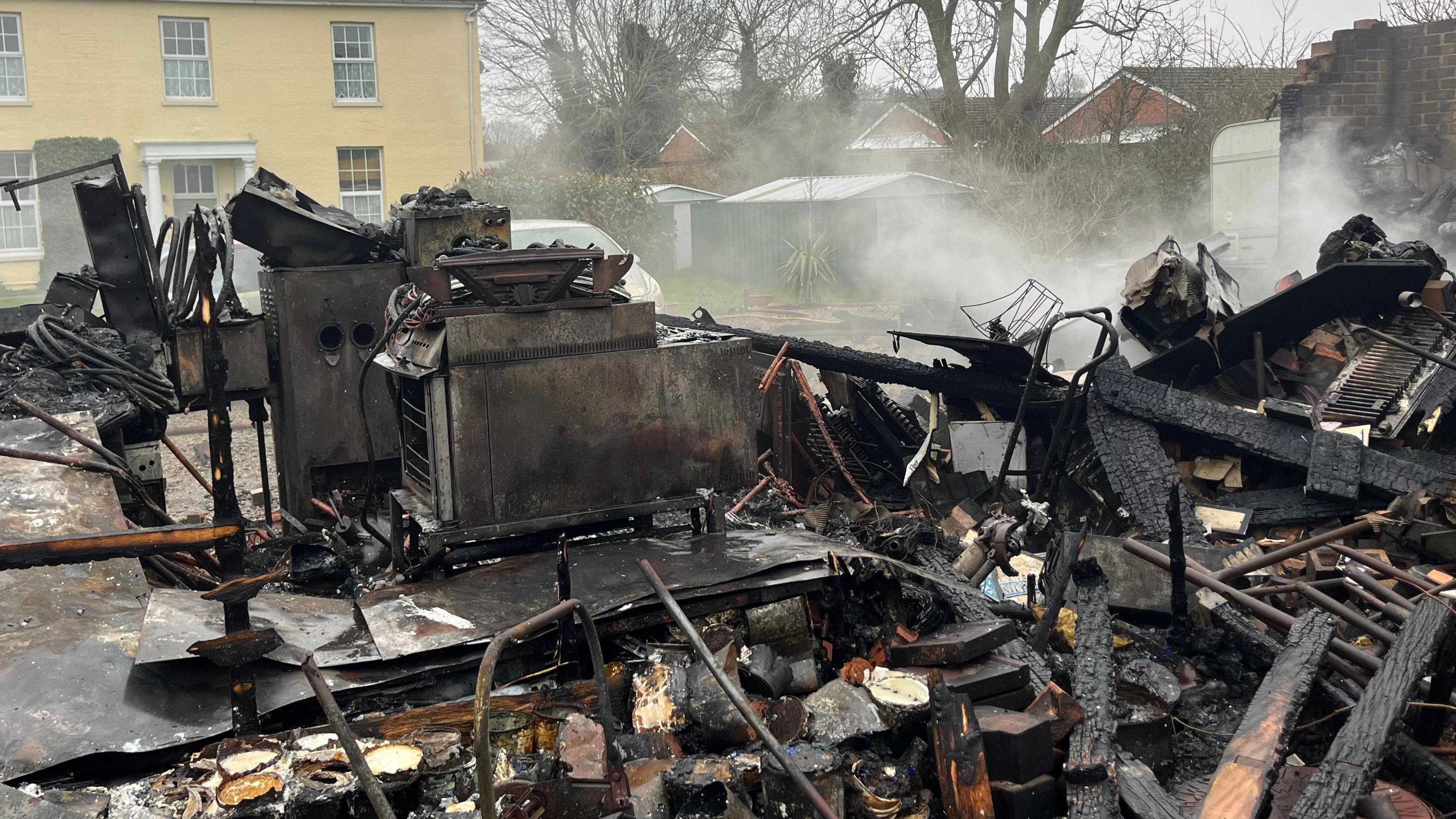 The wreckage of a fire outside a house. Burnt remains can be seen at the front of the building. Smoke can also be seen coming off of the wreckage. 