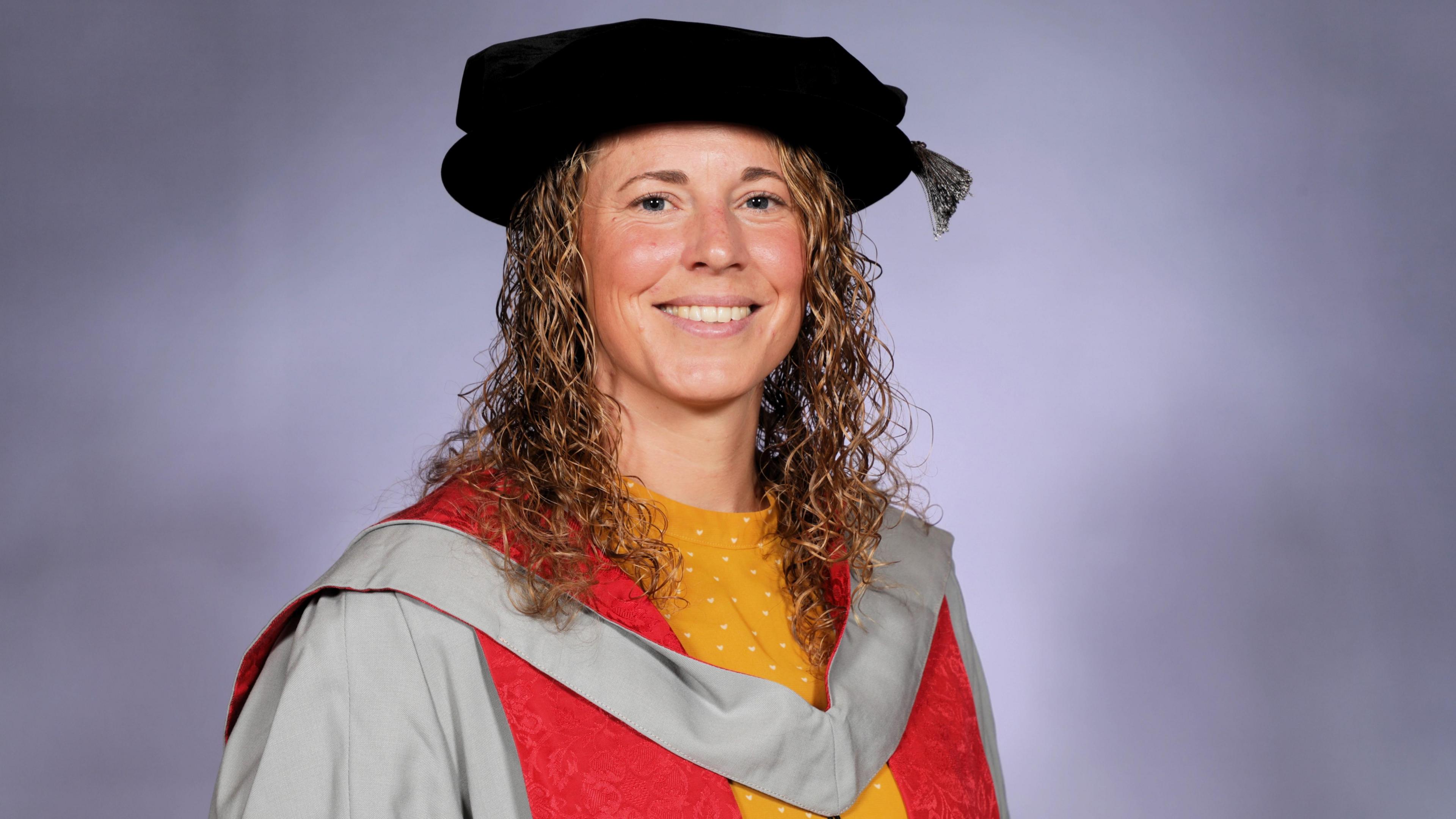 Amber Reed. She has blonde curly hair and is pictured wearing a graduation cap and gown. She is smiling at the camera. 