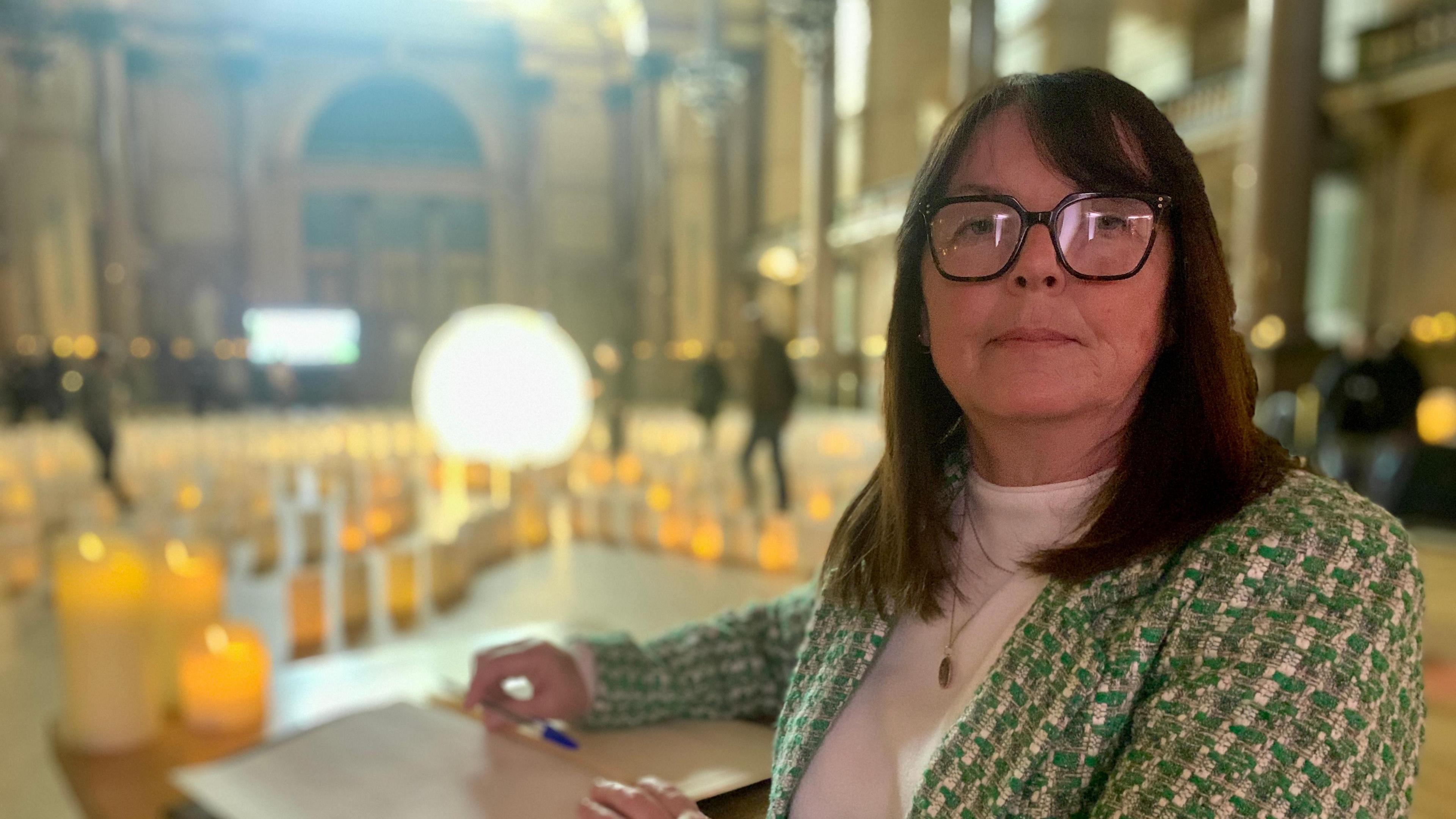 Barbara White stands beside the book with candles and paper lanterns glowing in the backdrop inside St George's Hall. She has shoulder-length brown hair and glasses and is wearing a green and white blazer.