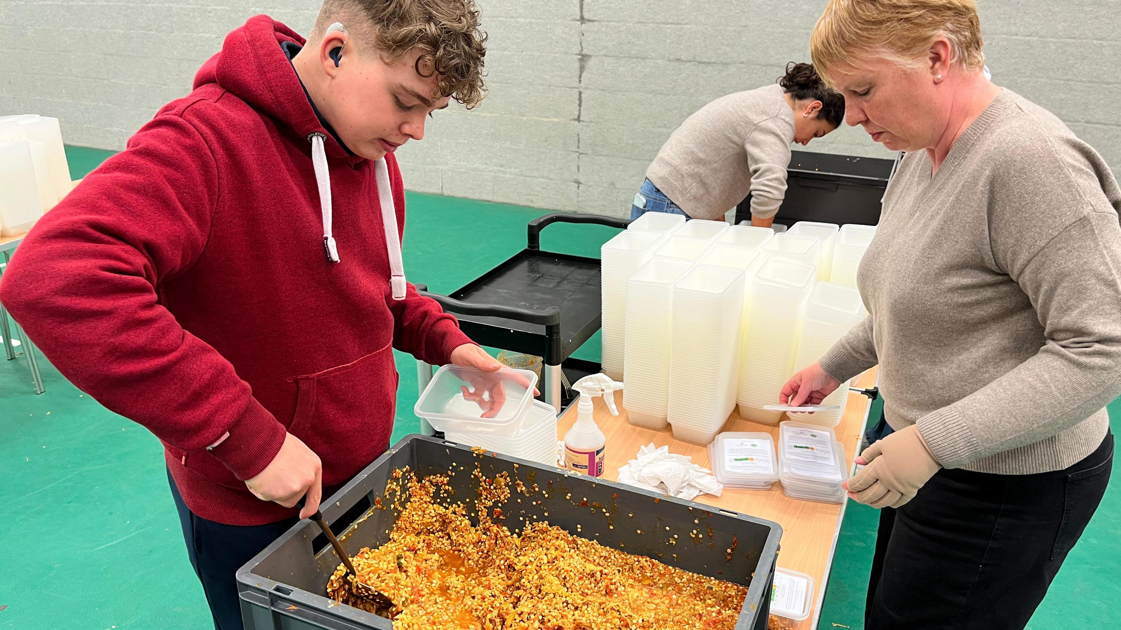 Two people filling plastic tubs with lentil dhal from a larger box. One the left is a teenage boy wearing a red hoody scooping the food into a tray, while on the right is a woman about to put a lid on a filled tub.