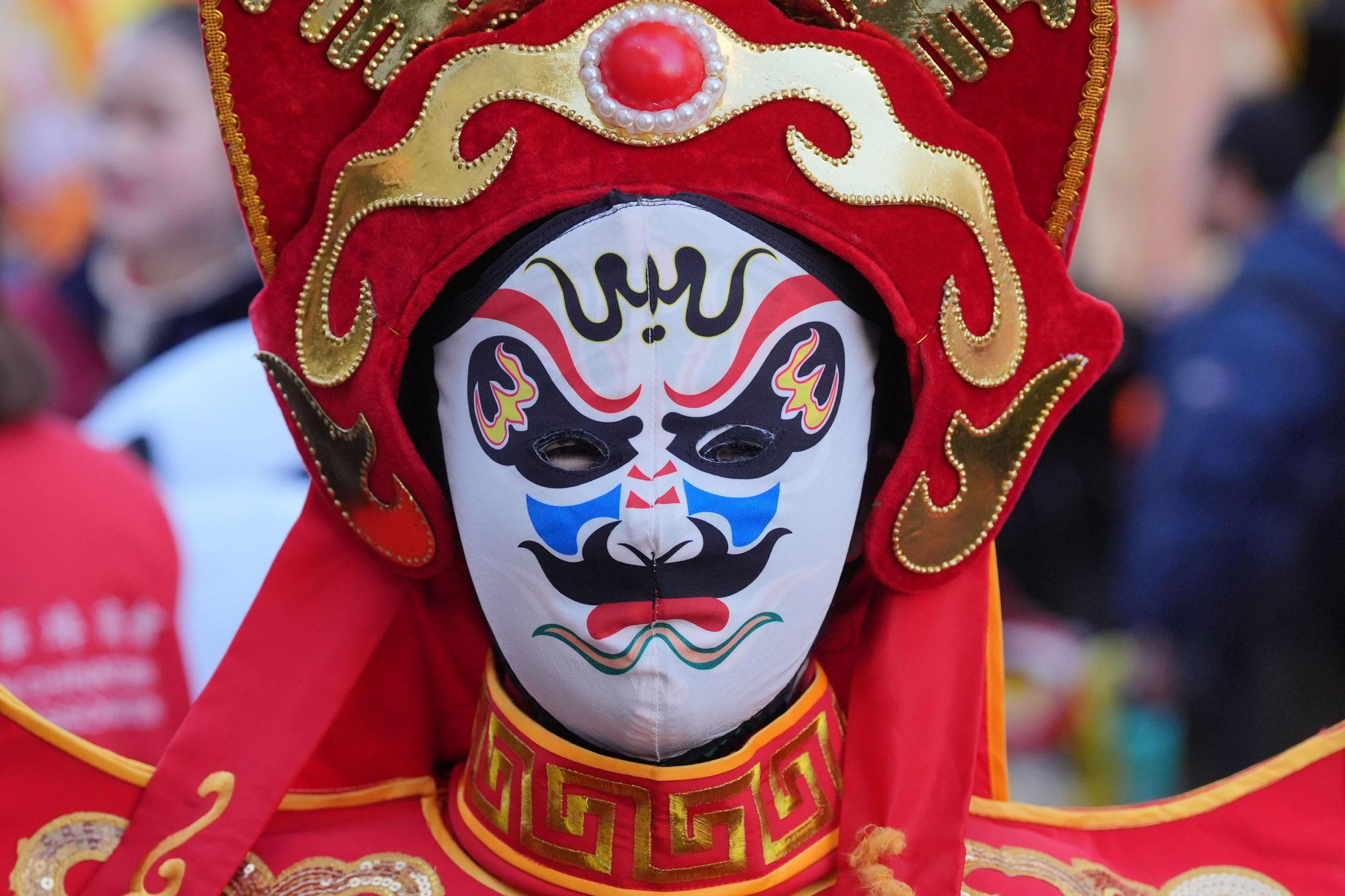 A performer is seen close up in a traditional Chinese costume wears a red and gold headdress and a white mask with intricate blue, red, and black painted designs. The mask has a fierce expression.