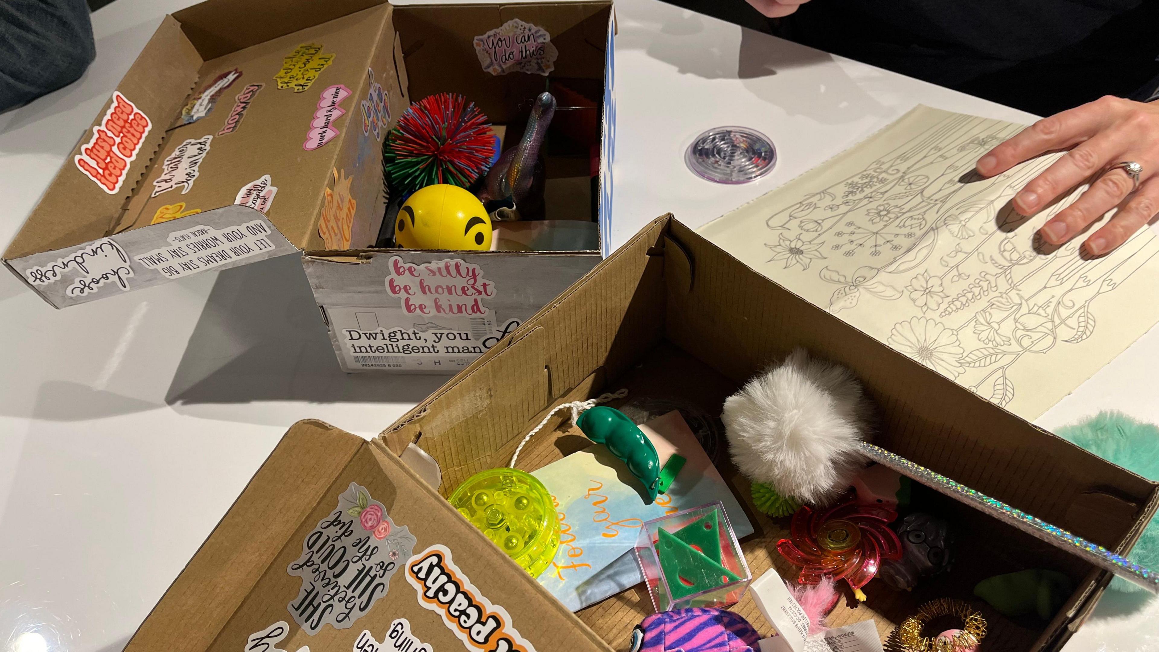 Two open shoeboxes with stickers on the outside showing positive messages. Inside are lots of fidget toys, stress balls, a diary and a pen with a fluffy top.