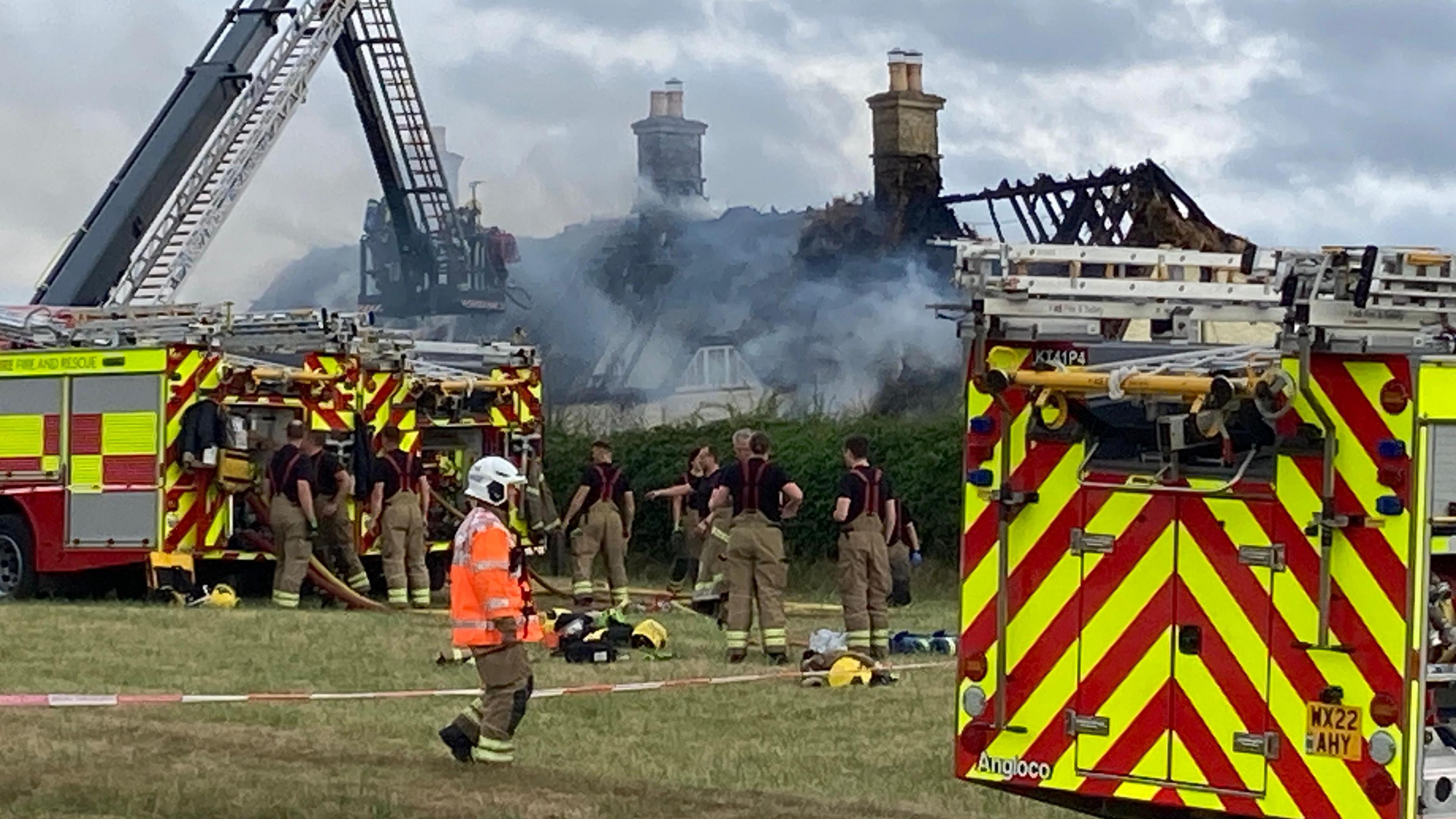 Three fire engines and an aerial ladder can be seen parked up on grass outside a cottage. About a dozen firefighters are working around the engines, with hoses coming out of them. The cottage in the background is smoking and the fire has left the bare structure of the roof on show.