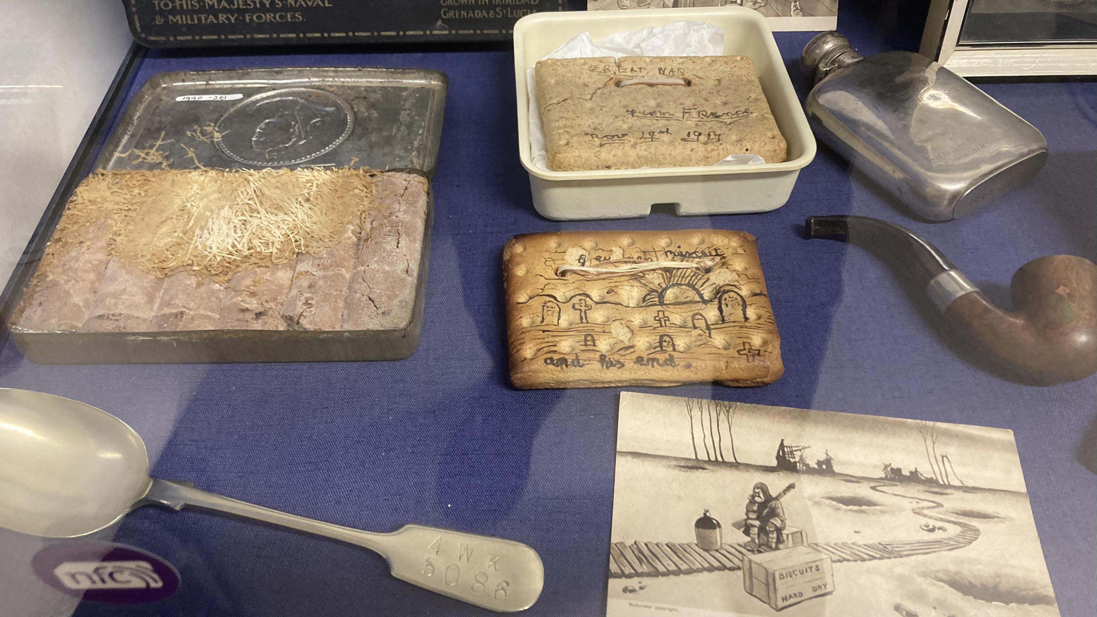 A beige biscuit with a drawing of a graveyard on it. There is a brown smoking pipe and a large discoloured silver spoon. There is another beige biscuit that reads great war