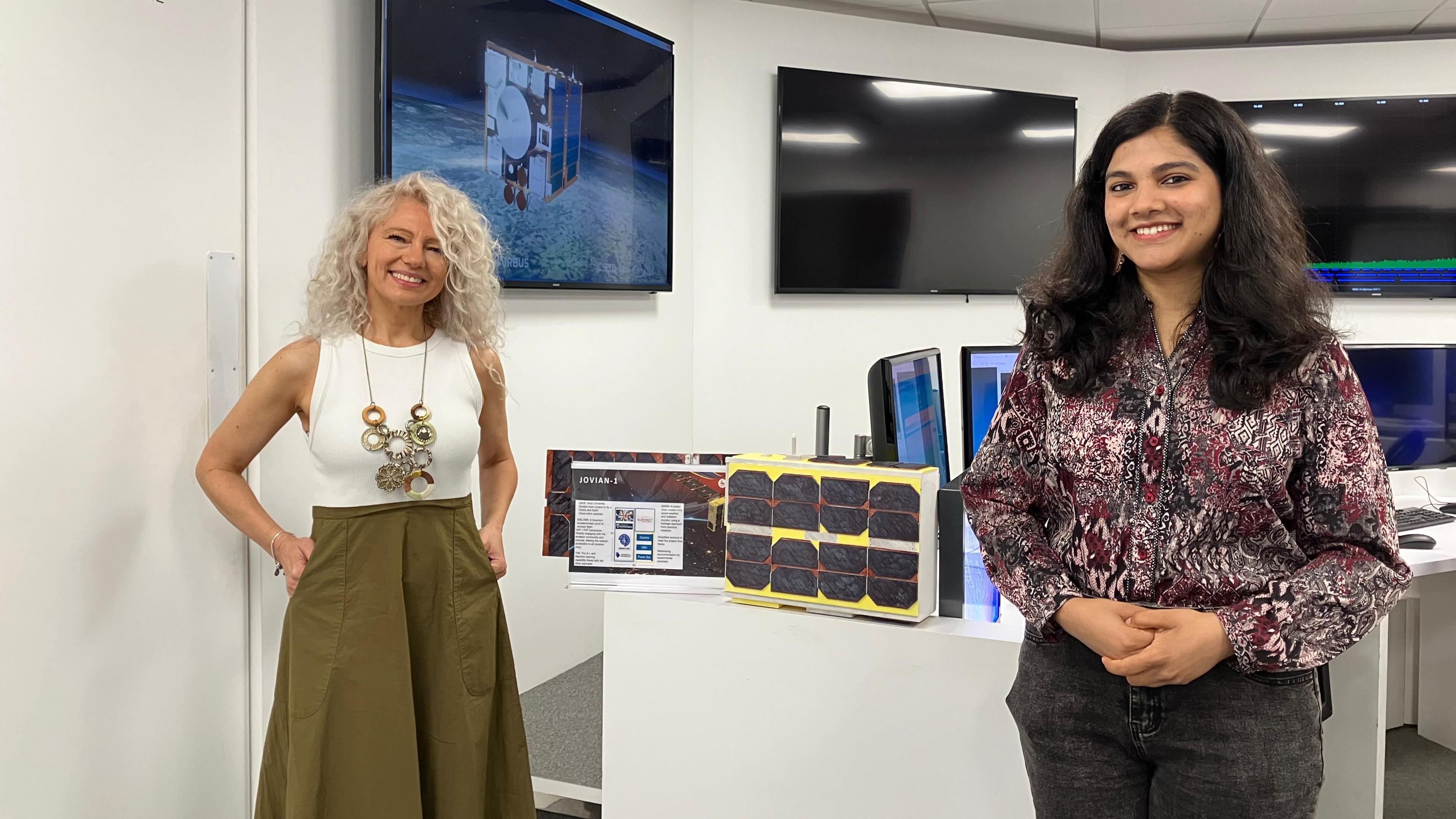 Two women stand either side of a small model satellite with black panels on the side and painted yellow. They are standing in front of computer and TV screens, one showing an image of a satellite in space.