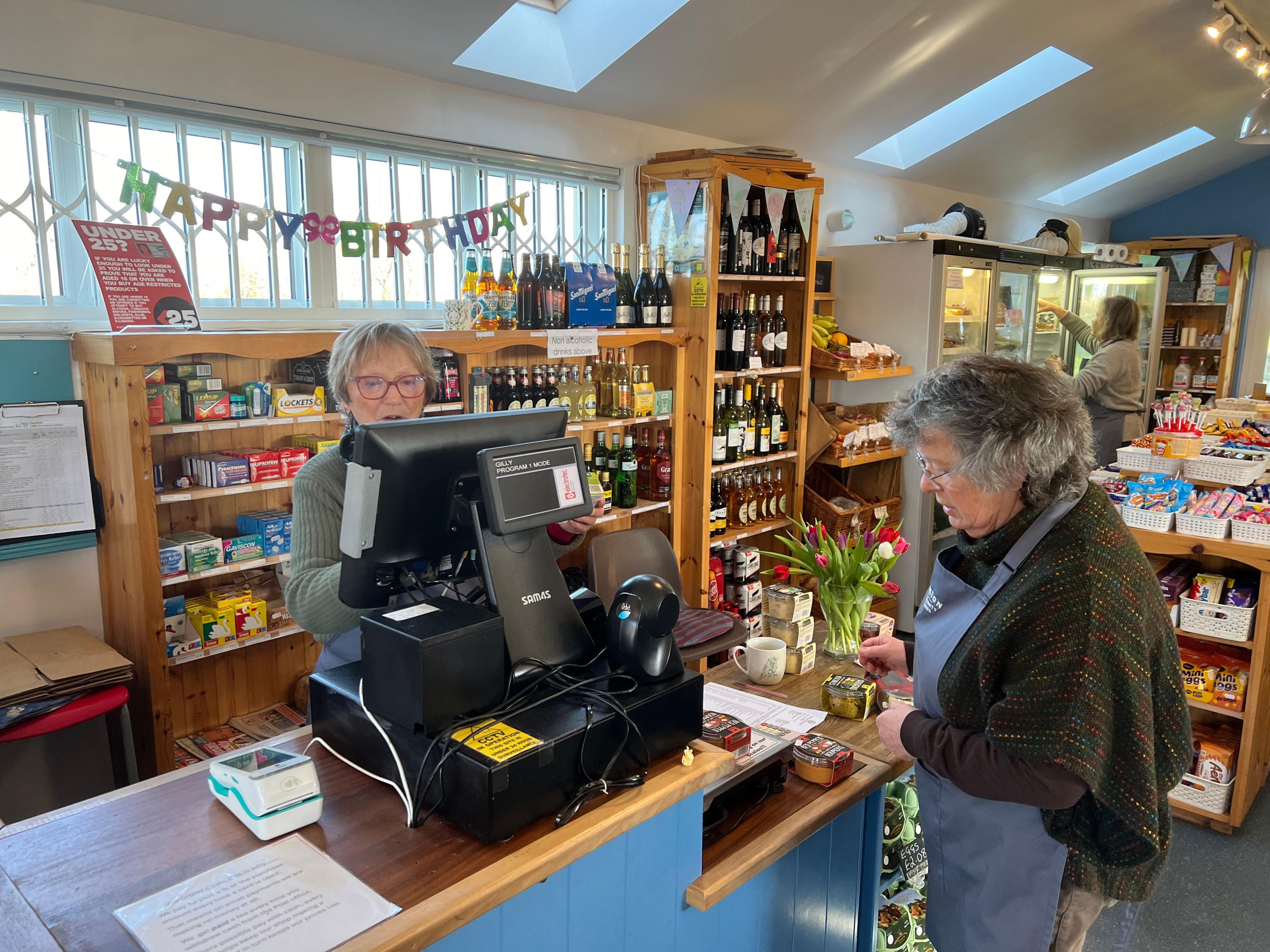 A lady standing behind a till in a well stocked shop serves a customer.