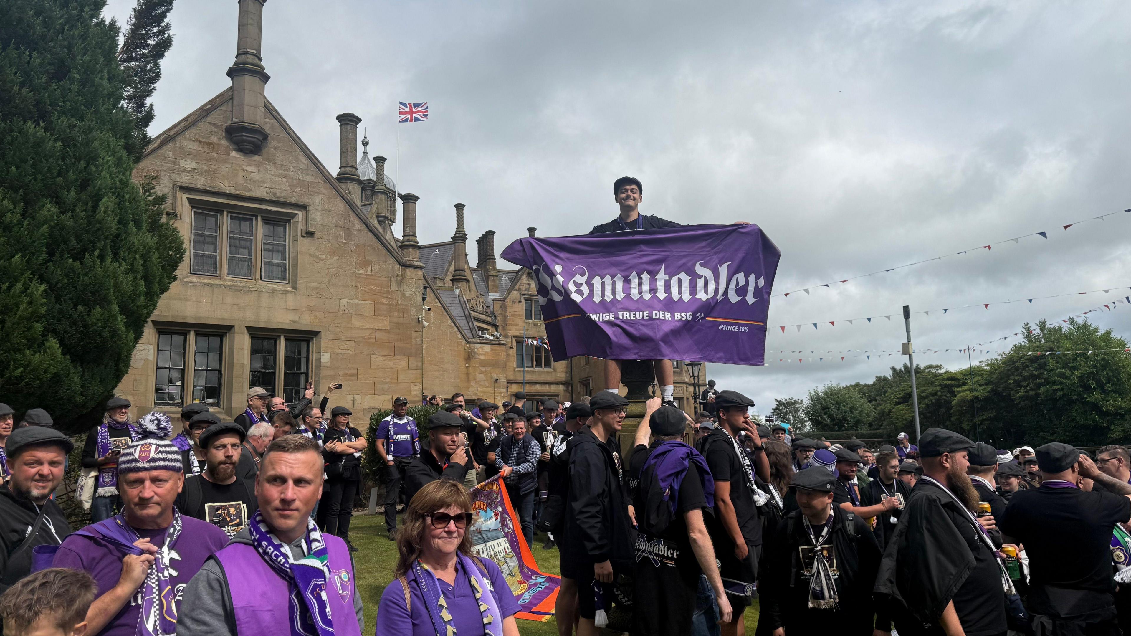 A large group of Erzgebirge Aue fans holding flags and banners standing outside Brownlow House - a brown brick building in a Baronial style