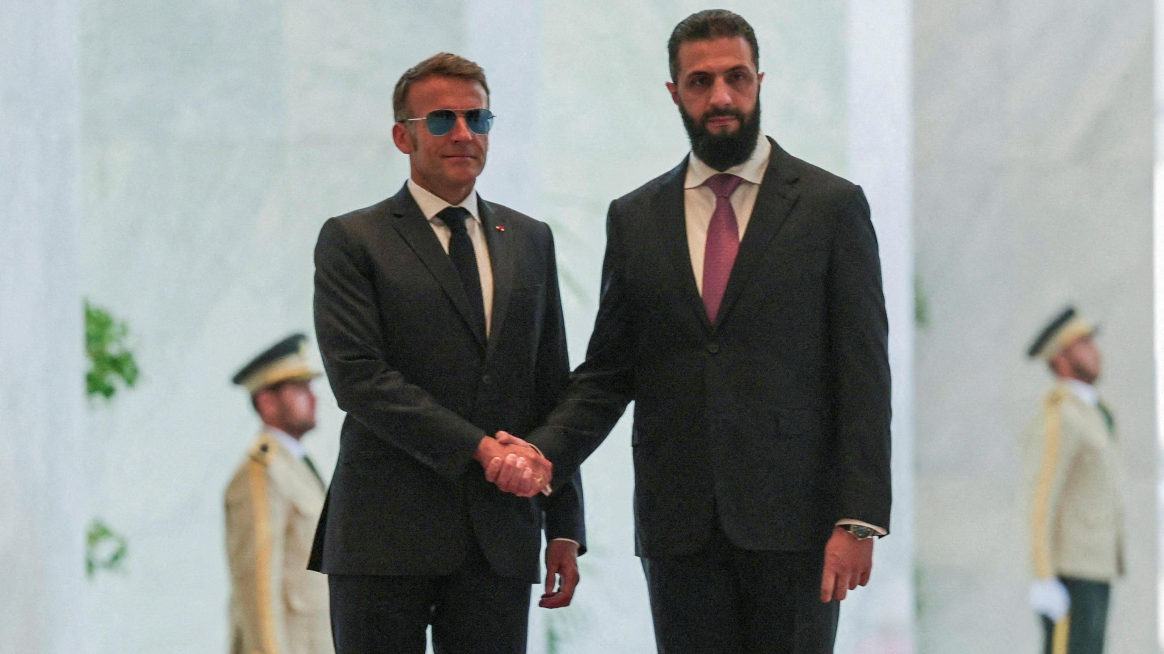 Macron wears a pair of sunglasses as he shakes hands with al-Sharaa