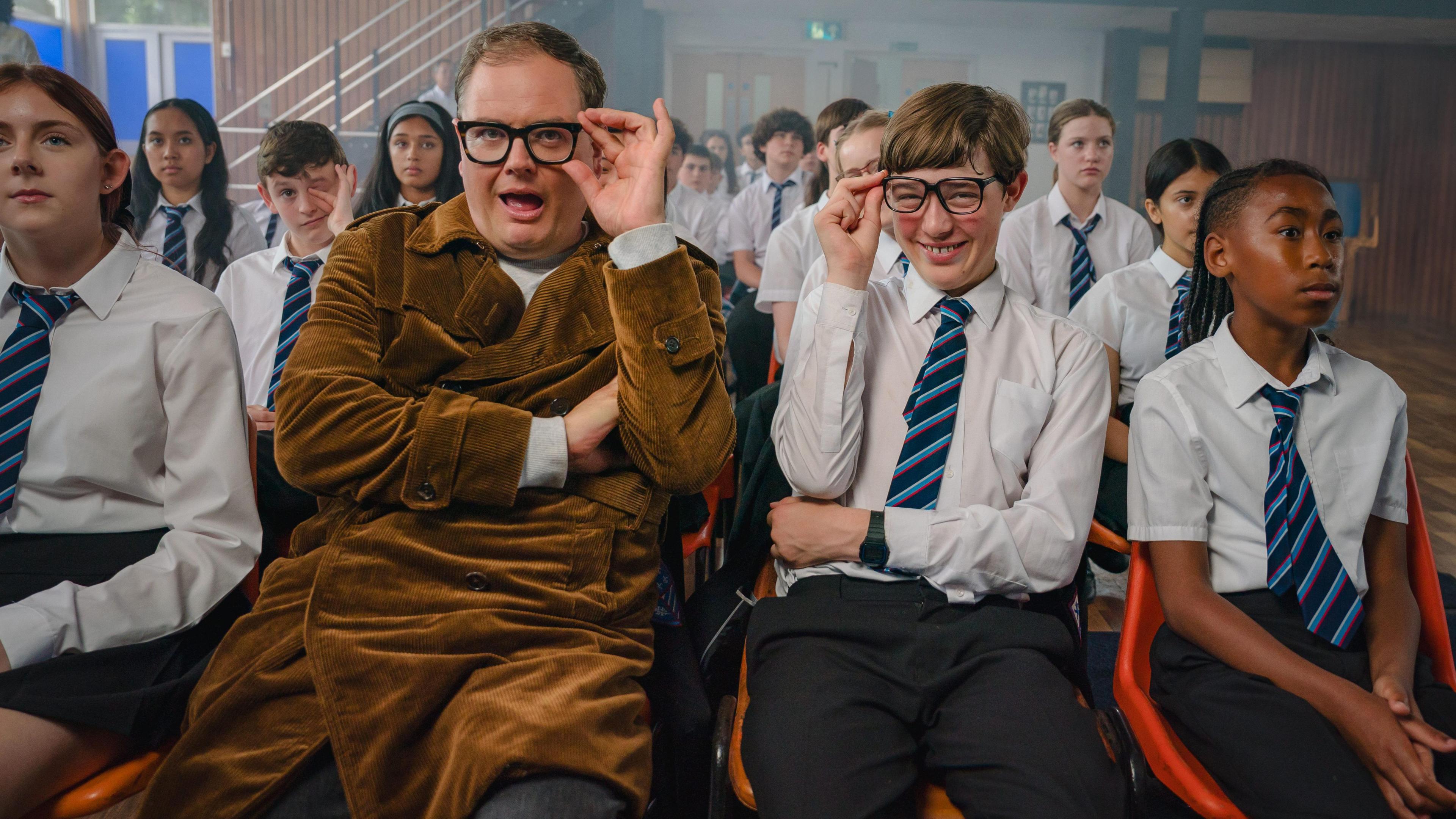 Alan Carr in a long brown coat holding his glasses and pulling a humour face. He is sat next to Oliver Savell who is a school uniform also holding his glasses and smiling. There are sat in a school hall among other pupils in school uniform.