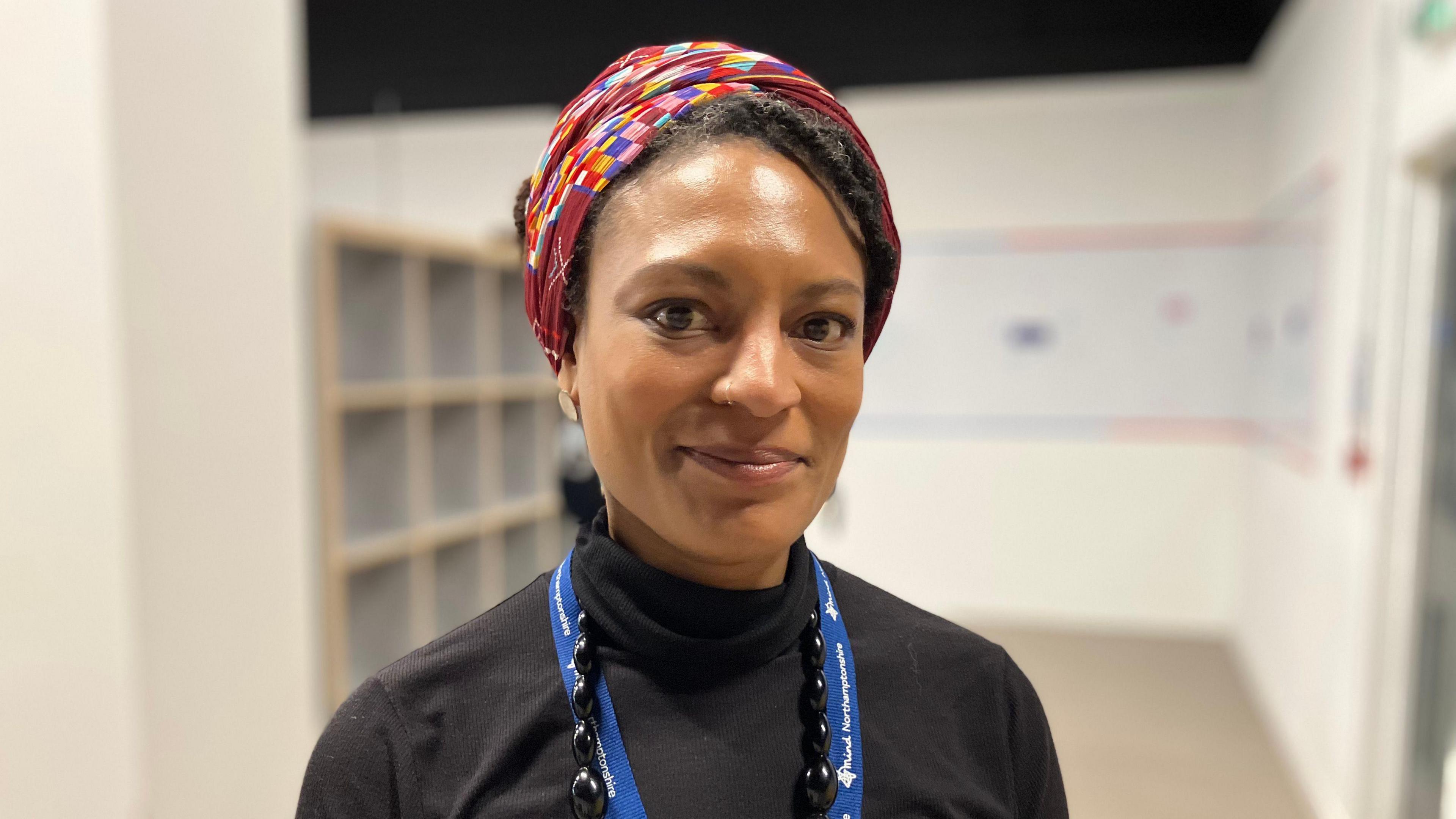 A woman in a black roll neck jumper, a blue lanyard, and a multi-coloured hair cover smiles without teeth at the camera.