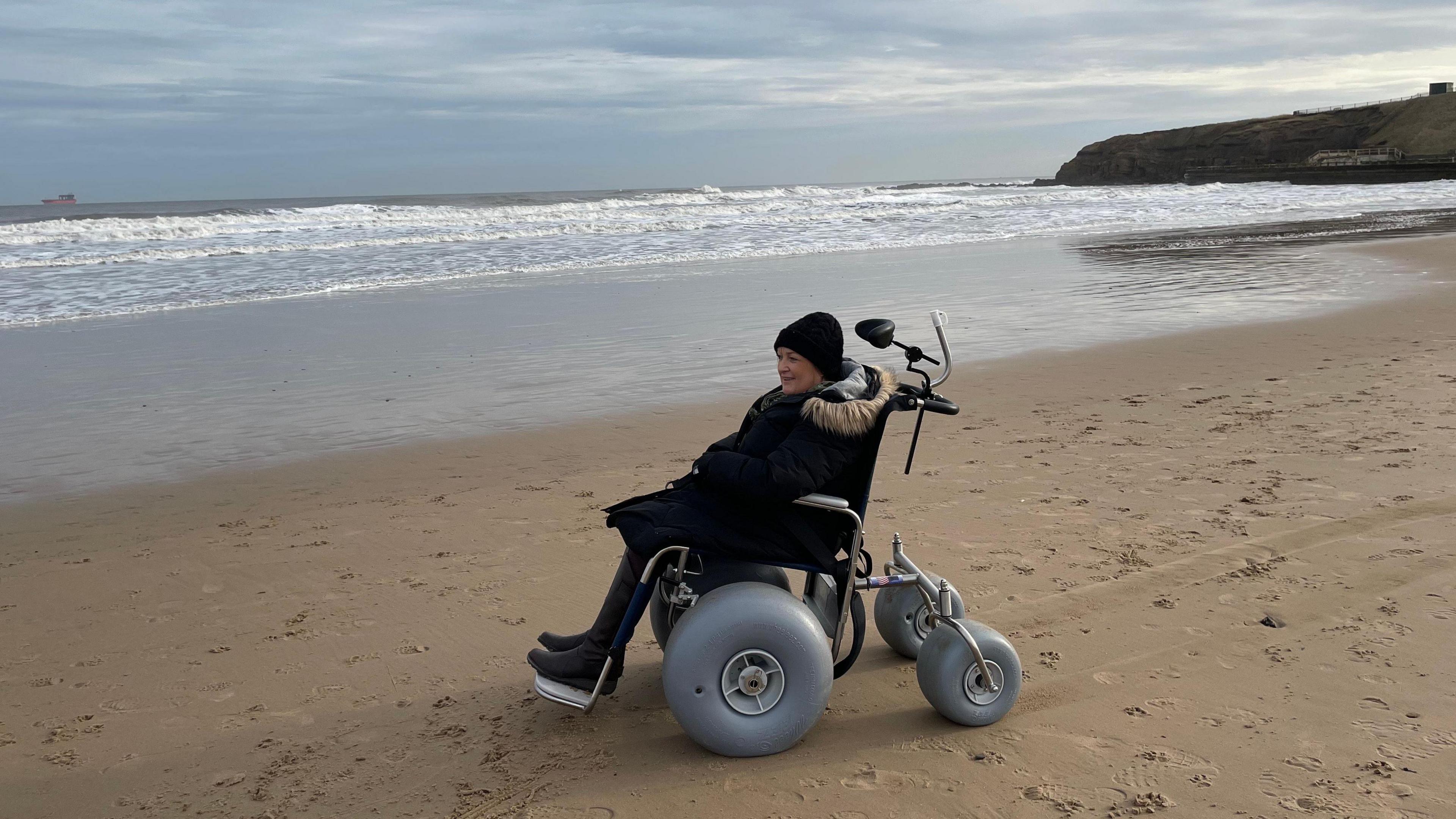 Woman sitting in a wheelchair on a beach, next to the sea shore. She is wearing a dark coat with a fur trimmed hood, black boots and a black beanie type hat. The wheelchair is one specially adapted for the beach as it has larger wheels than usual.