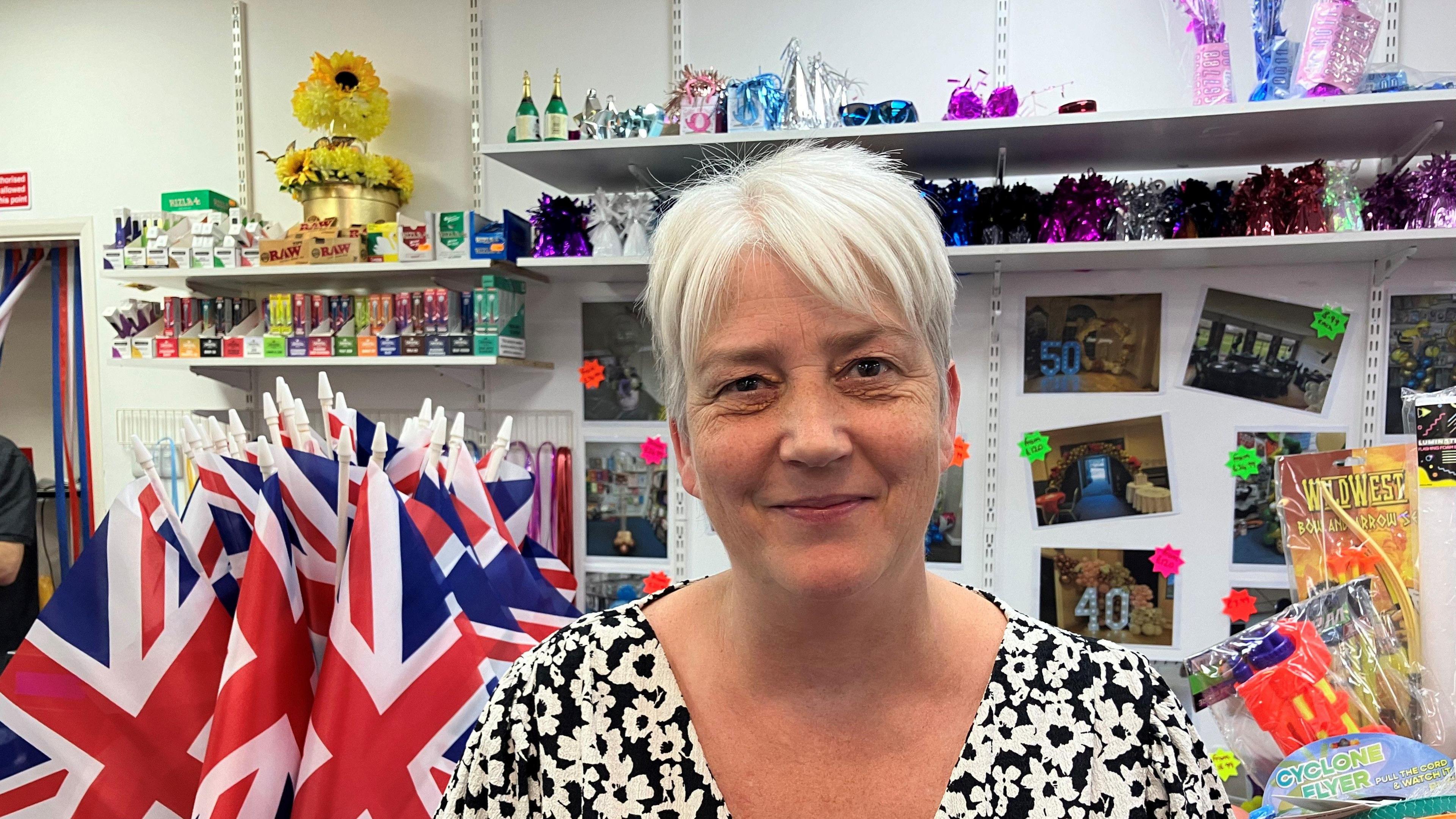 A woman wearing and black top with white flowers smiling, with short grey hair. She is standing in front of a shop shelf where there are Union jack flags, postcards and assorted party items in the background.