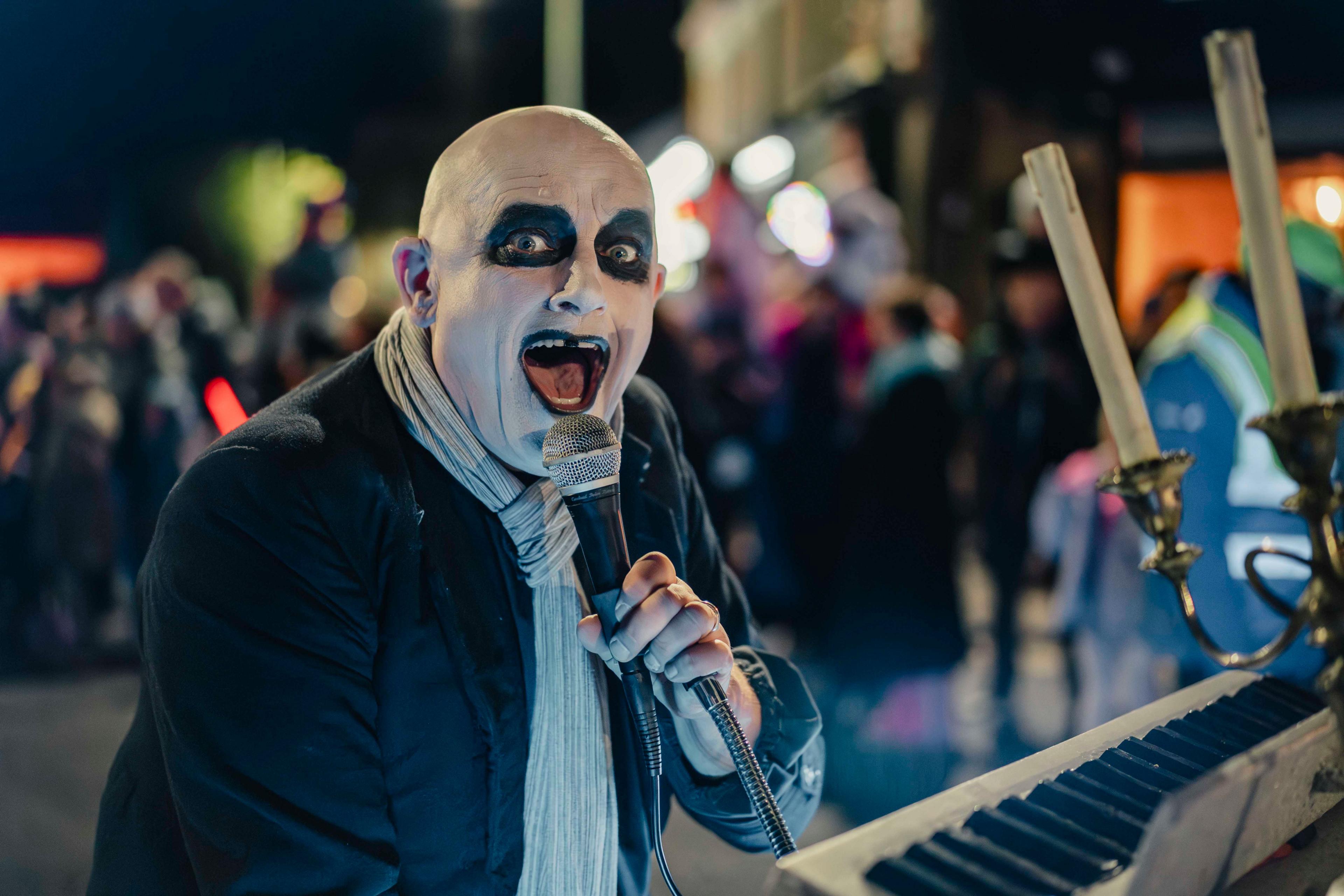A bald man in white face paint with black circles around his eyes stares into the camera. He is wearing a black blazer and a white scarf tied tightly around his neck. He sings into a microphone while playing an electric organ.