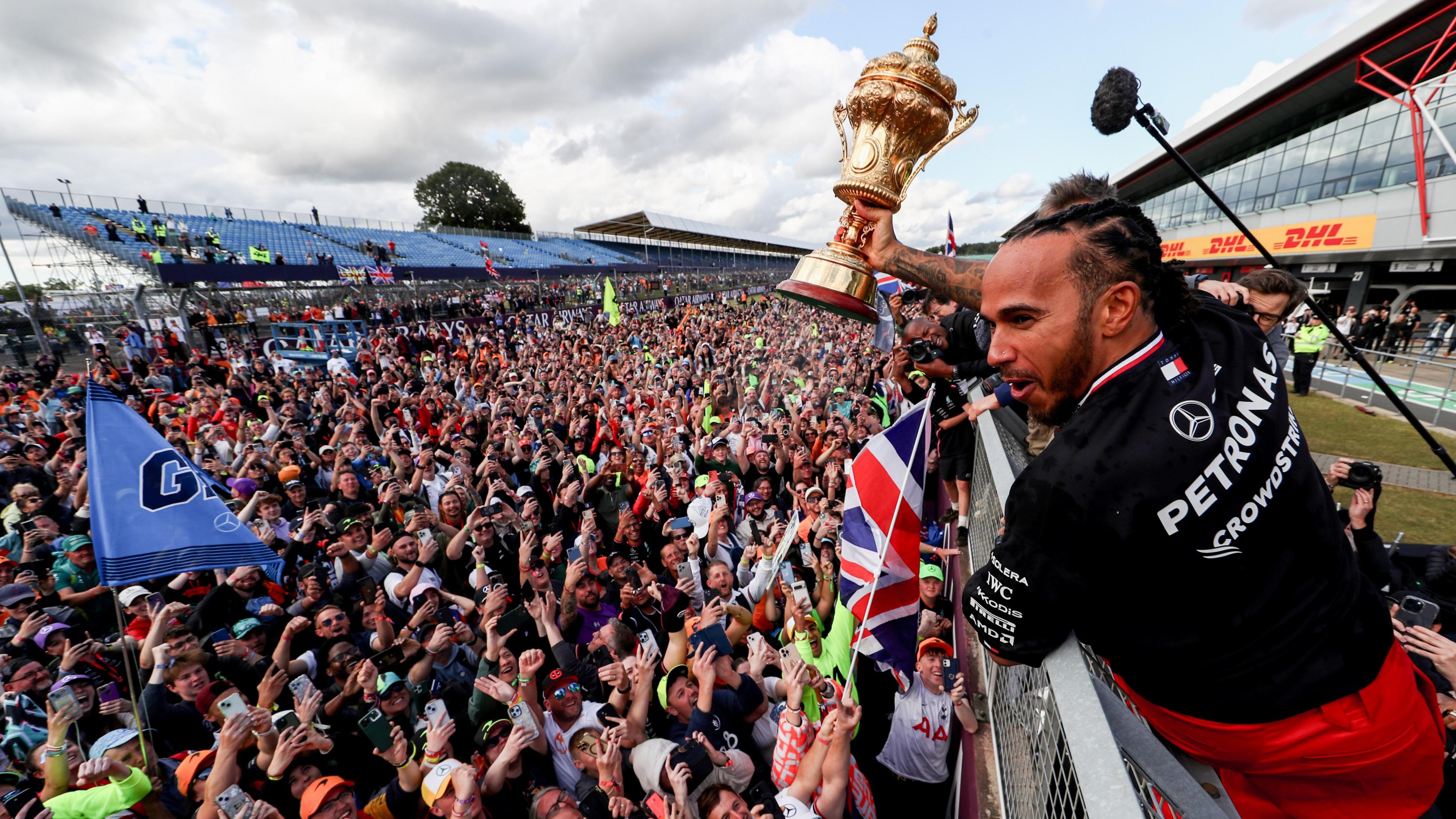 Lewis Hamilton holds a gold-coloured trophy aloft and smiles as a crowd looks on below him. Many are holding their phones up and there are several flags among them.