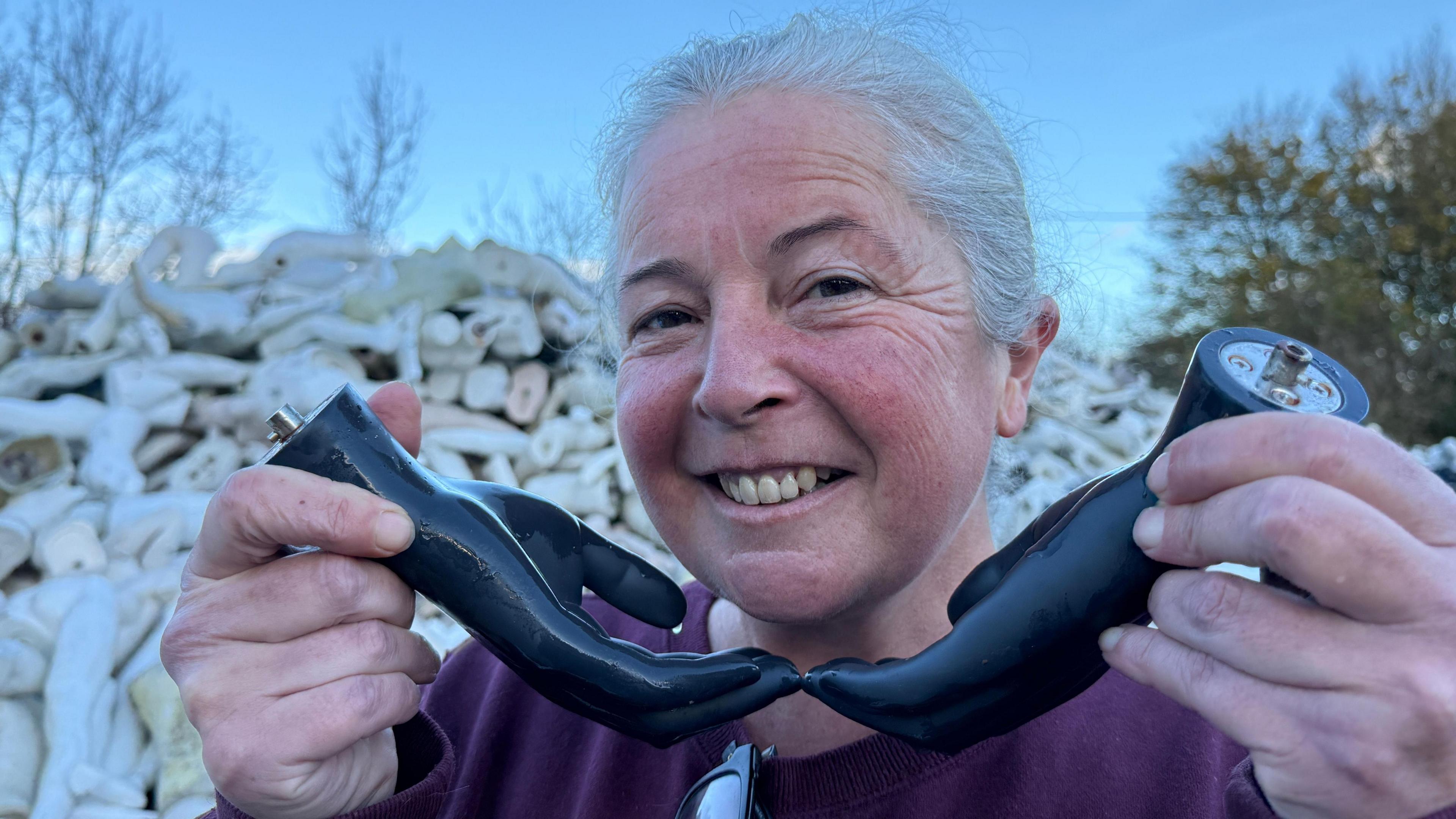 Roz Edwards is standing behind a mound of mannequin body parts. Legs, torsos, heads and feet are all packed together. She smiles, looking at the camera whilst holding a pair of mannequin hands, framing her face. 